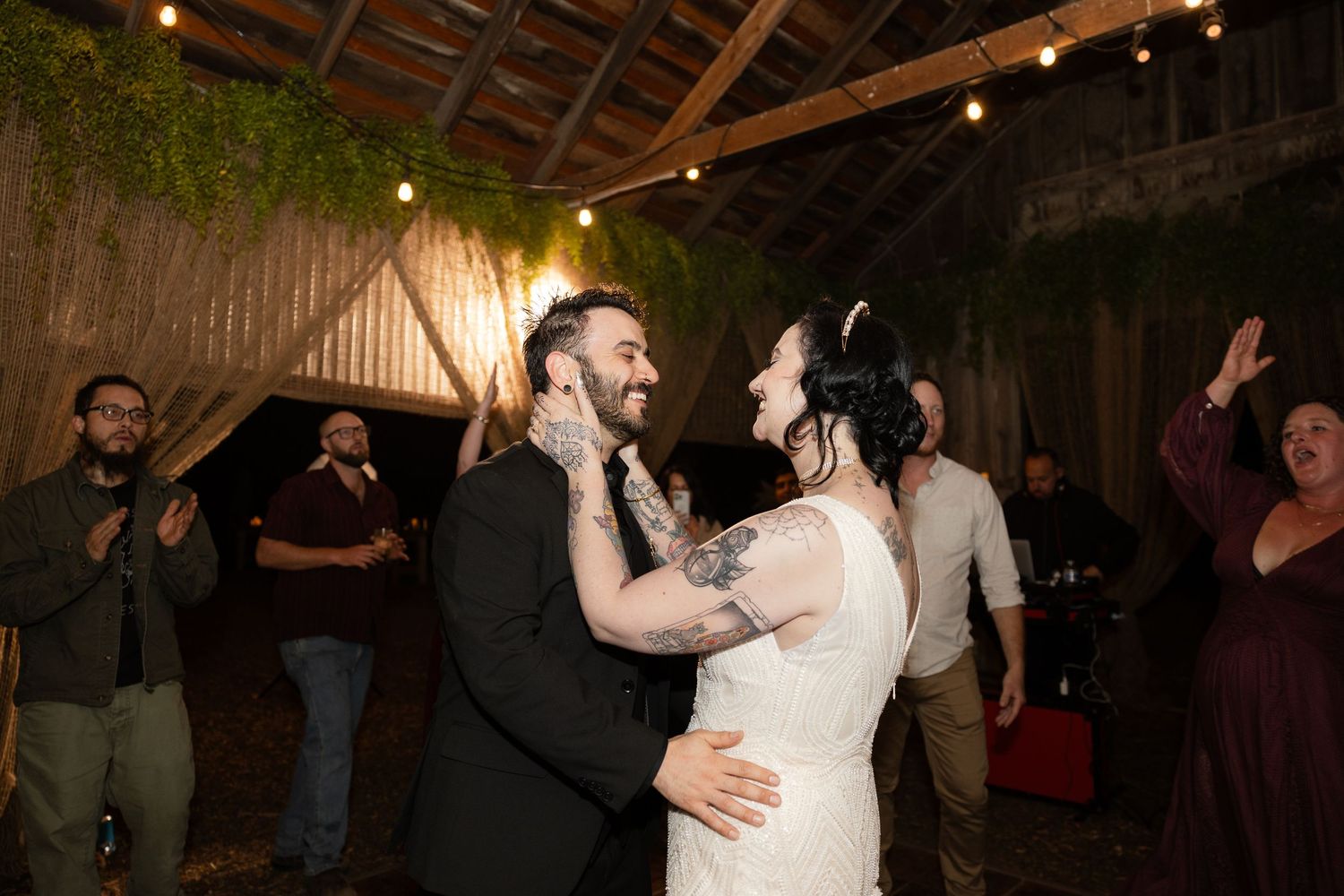 Wedding guests dance together during a lively reception in a rustic barn venue with string lights overhead.