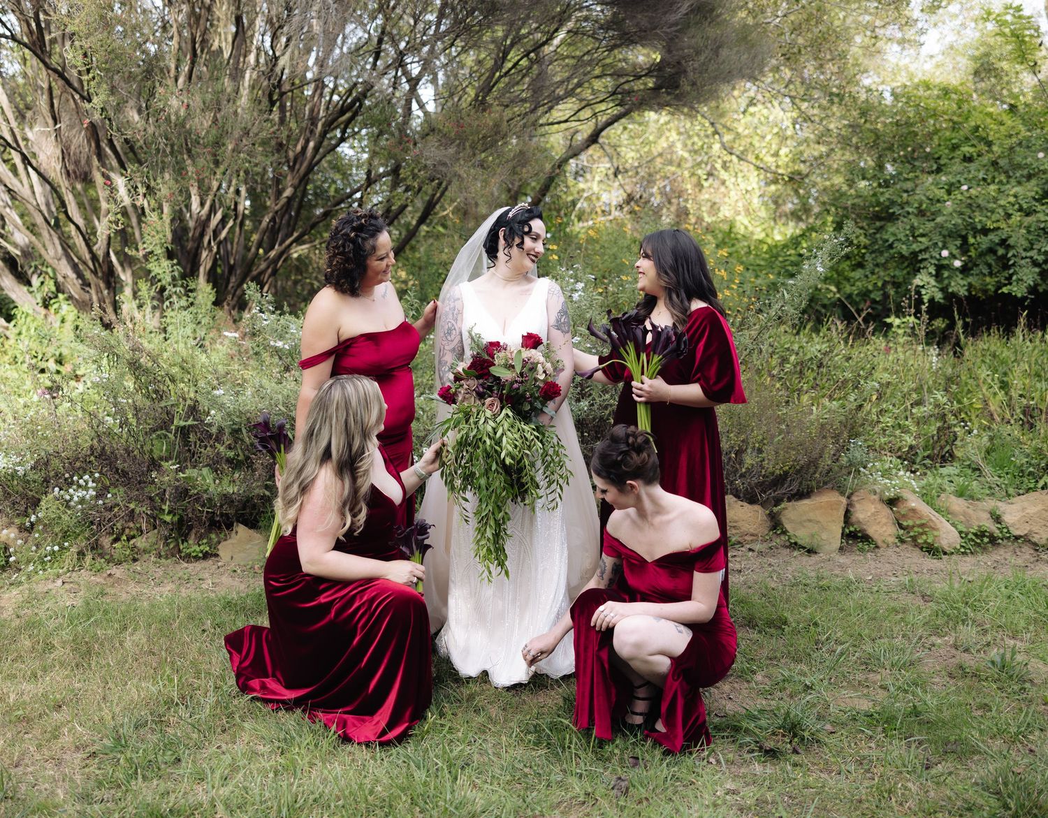 Bridesmaids in deep red dresses surround bride during outdoor photo session.