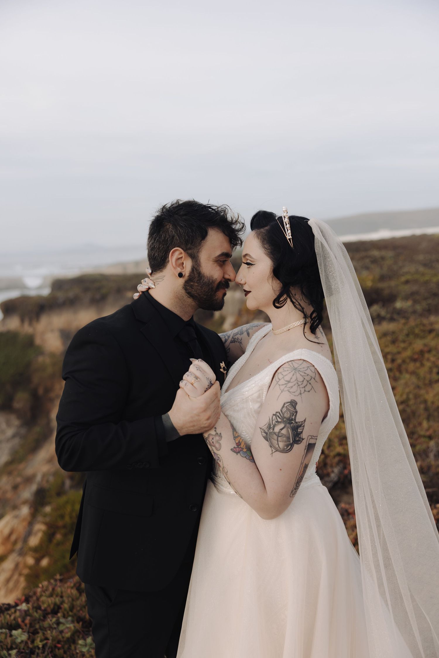 A romantic wedding couple shares an intimate moment on a coastal cliff with dramatic ocean views in the background.