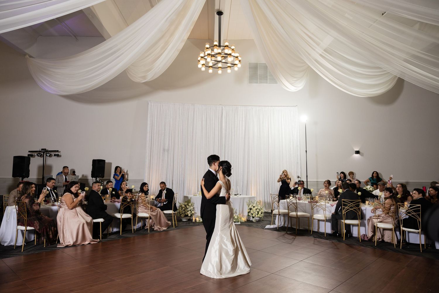 Elegant wedding venue with white fabric ceiling draping and modern chandeliers during reception.