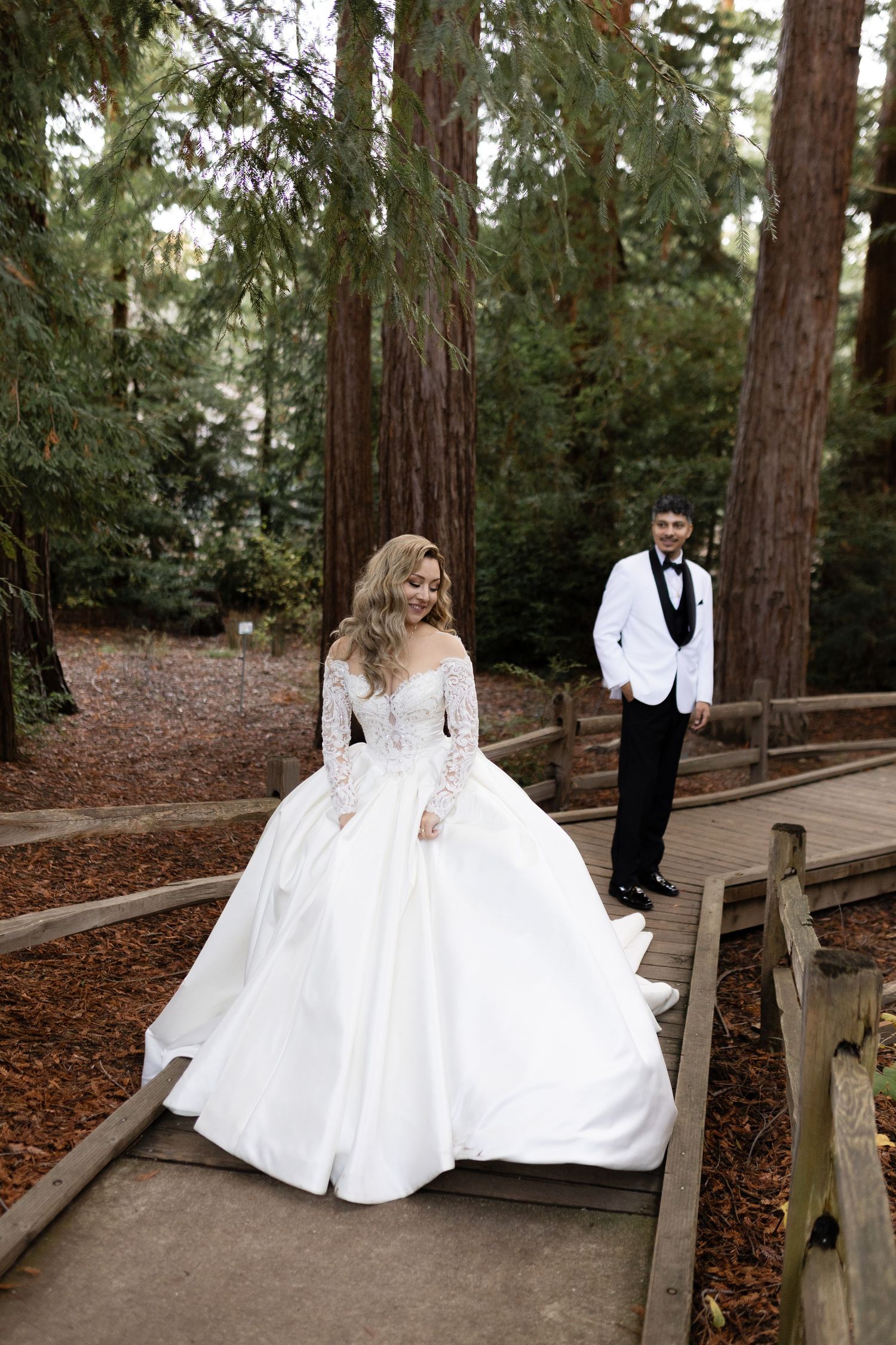 A bride in a luxurious white gown stands on a forest boardwalk with her groom in the background.