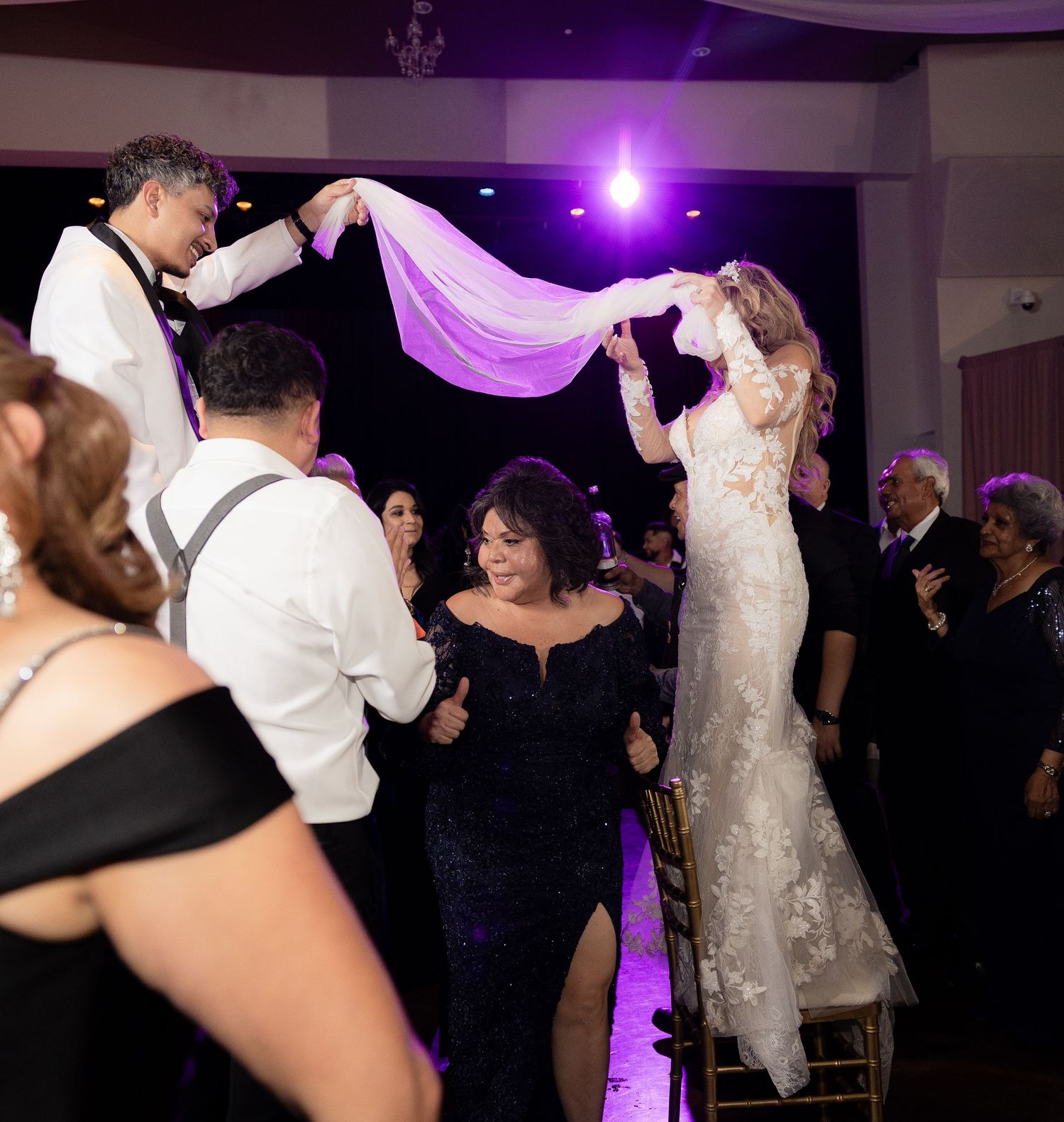 Wedding celebration with bride in white dress dancing among guests under purple mood lighting.