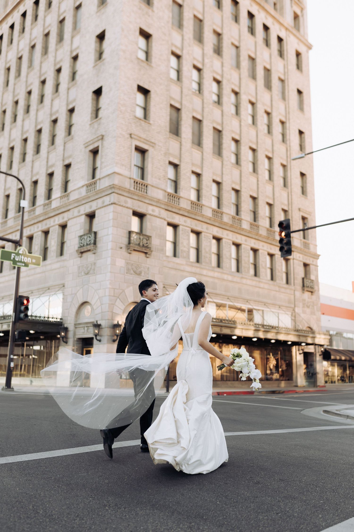 Couple in wedding attire walking past historic downtown building with flowing veil in the wind.