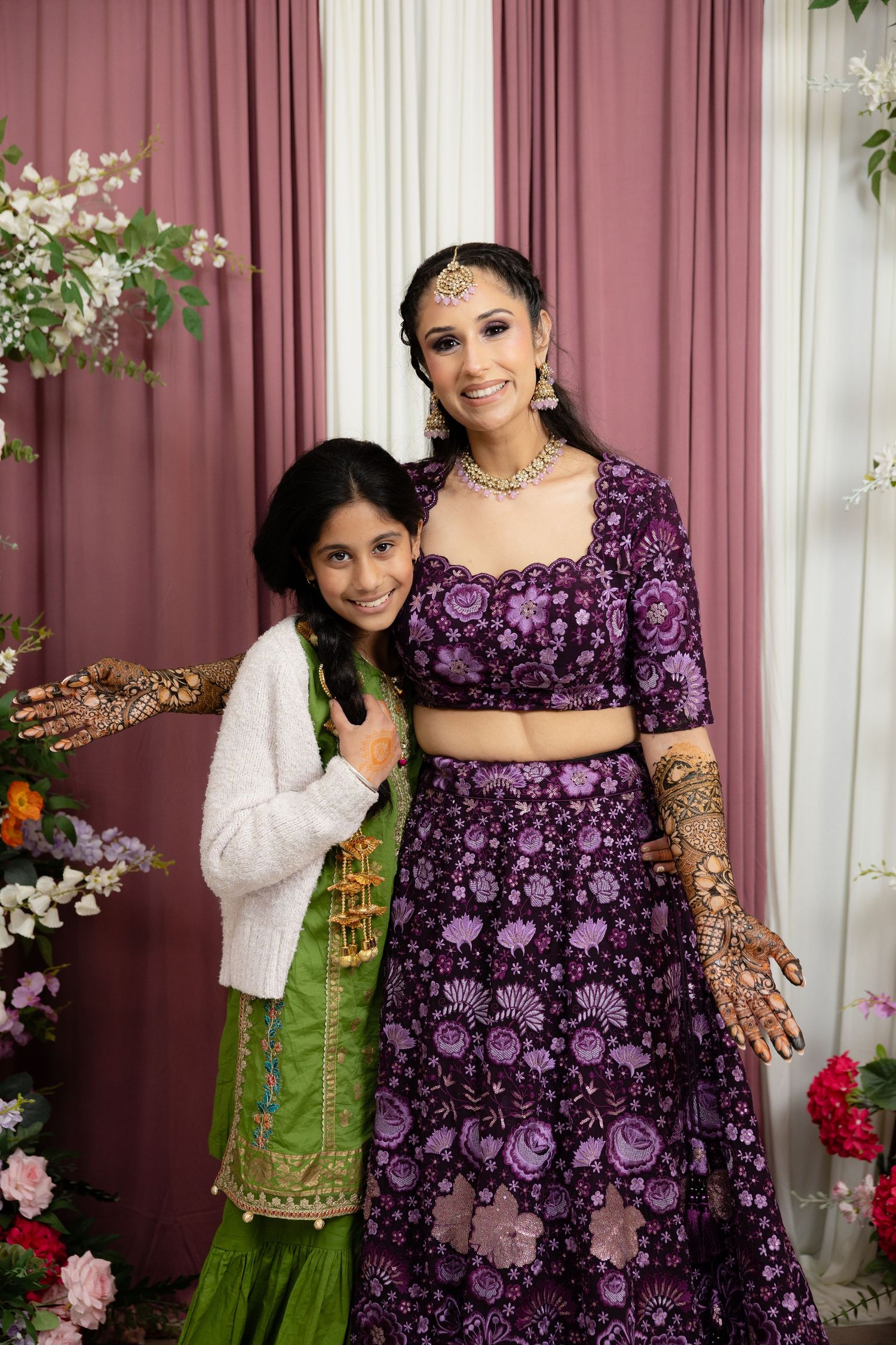 A group poses together wearing traditional South Asian attire including purple and green garments with henna designs.