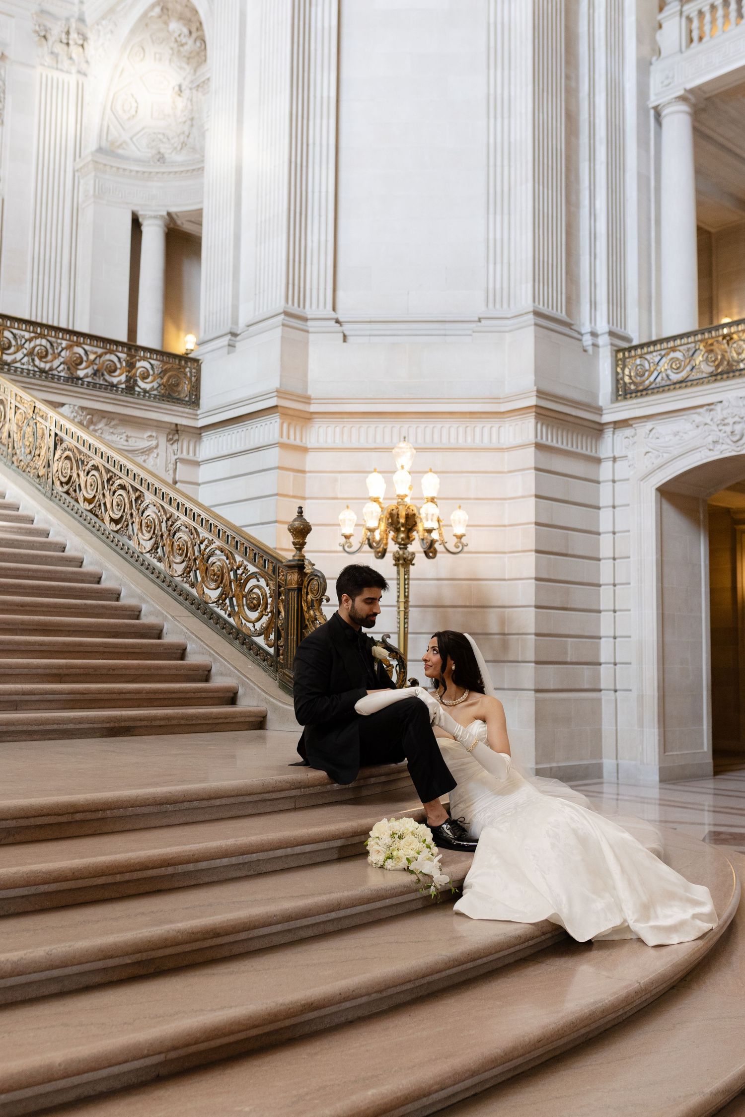 A couple sits on grand marble stairs with ornate railings inside a luxurious historic building.