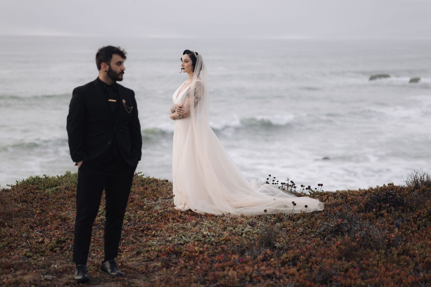 Wide landscape shot of couple on rugged coastline with waves crashing below.
