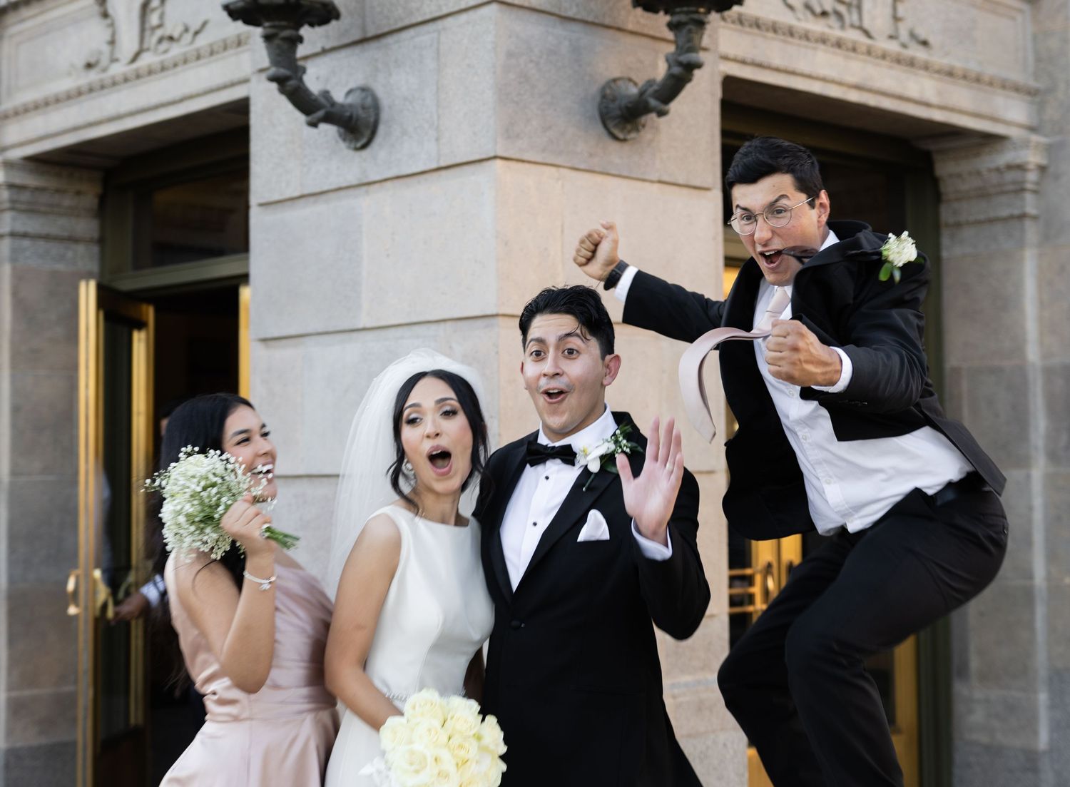 Wedding party members strike playful poses outside stone building entrance.