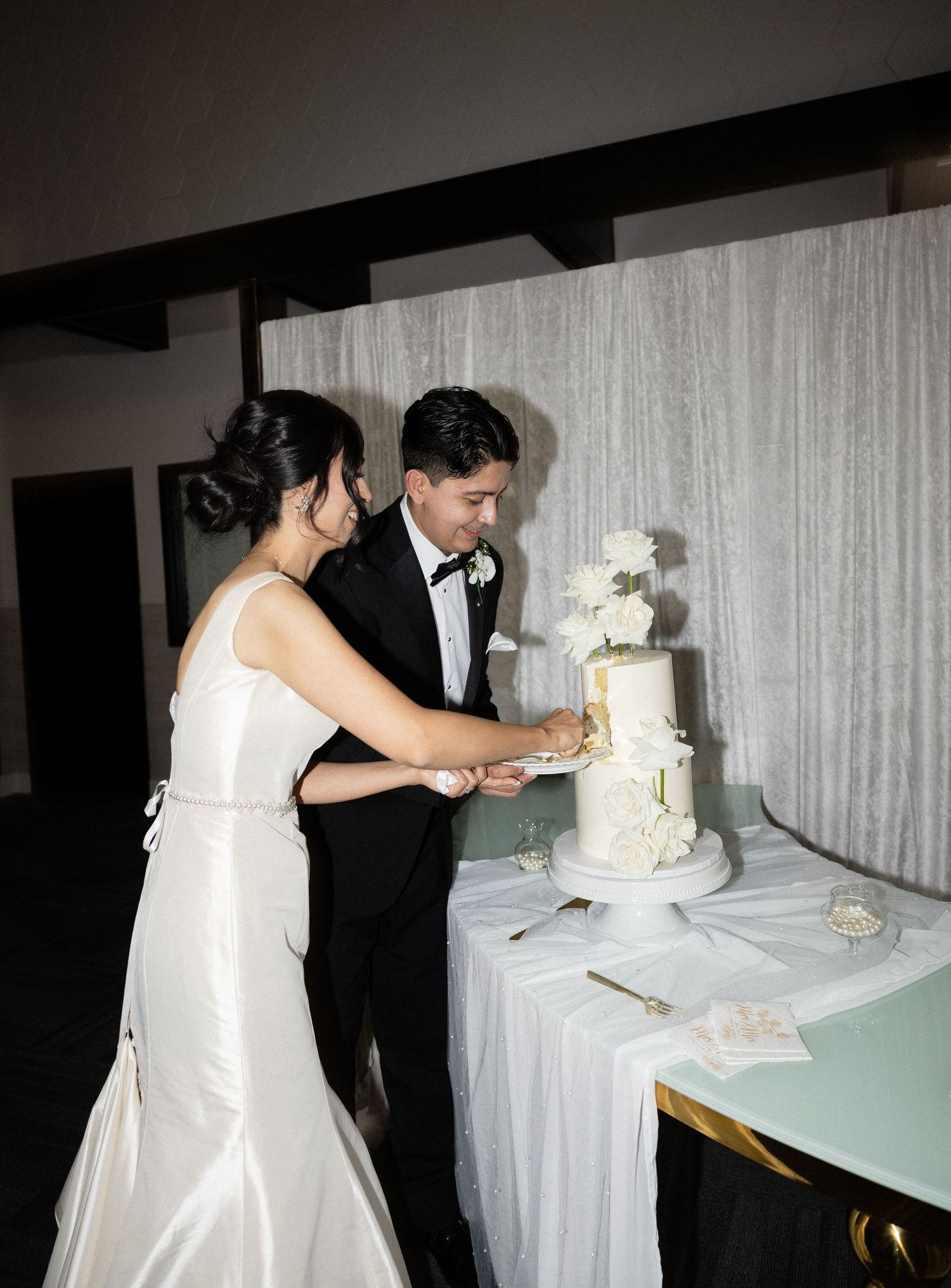 Wedding couple shares a fun cake-cutting moment at their reception with a white-draped backdrop.