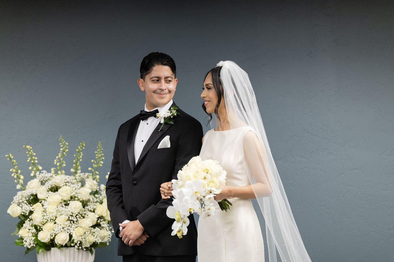 Elegant wedding portrait with white floral arrangements against grey studio backdrop.