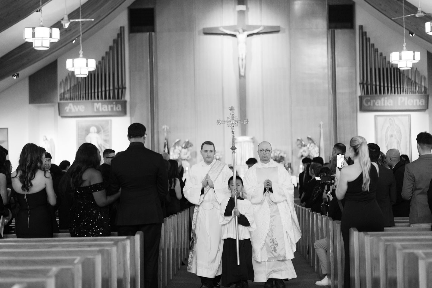 A black and white photo of a church processional with clergy and attendants walking down the aisle.