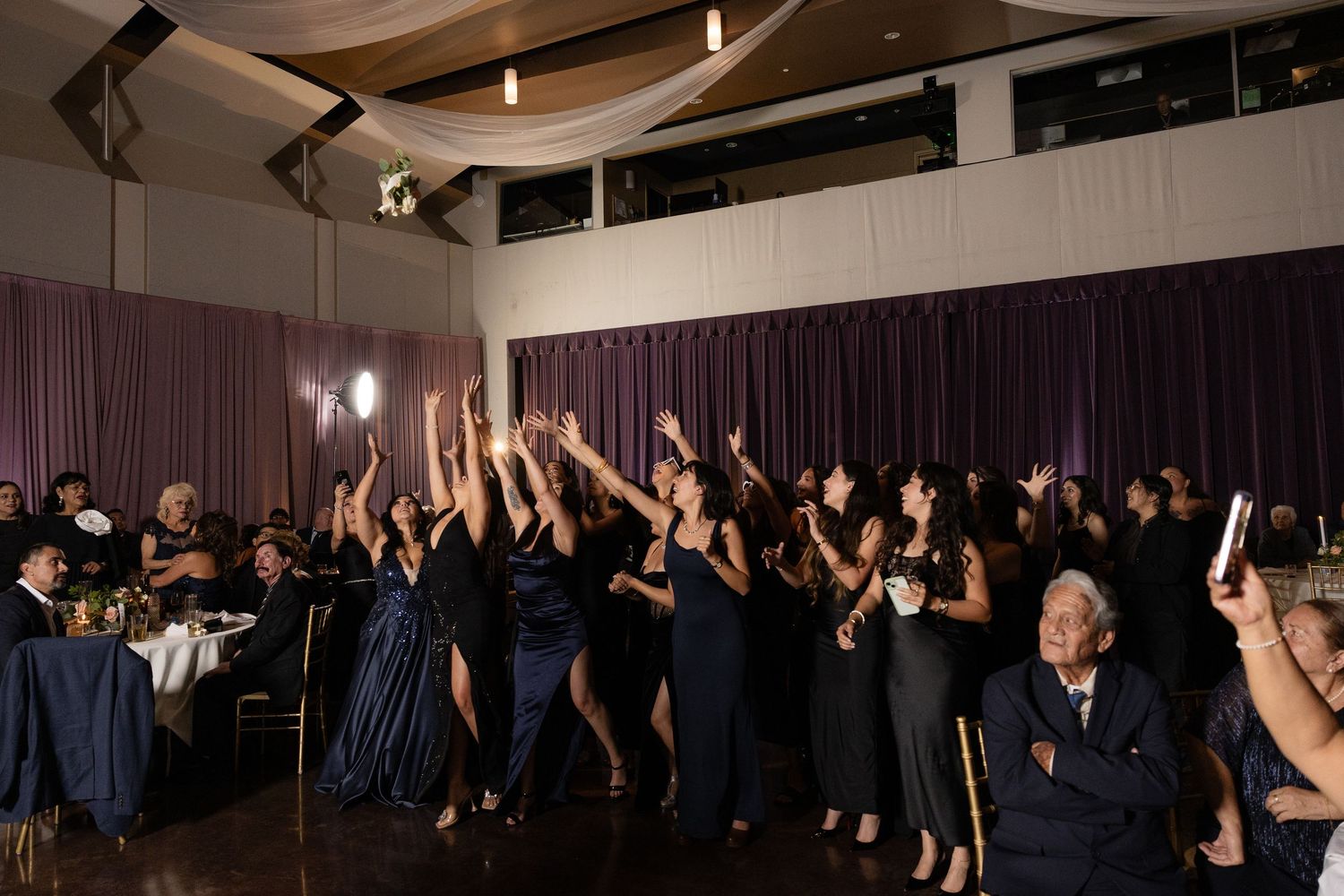Large group of wedding guests dancing together in a dark reception venue.