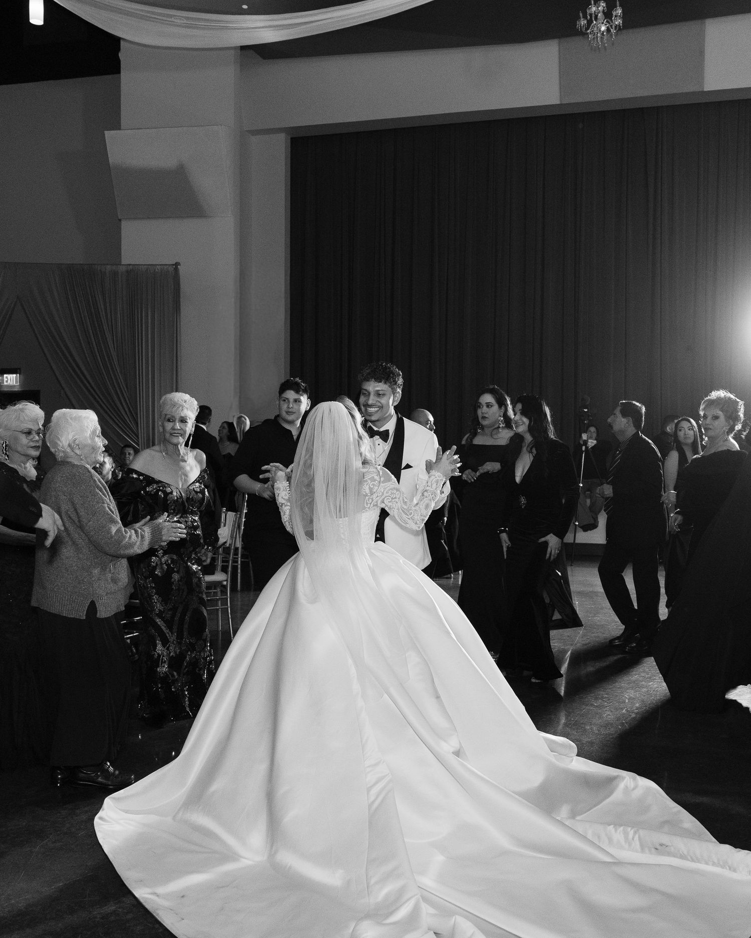 Black and white photo captures wedding couple's first dance surrounded by guests at reception.