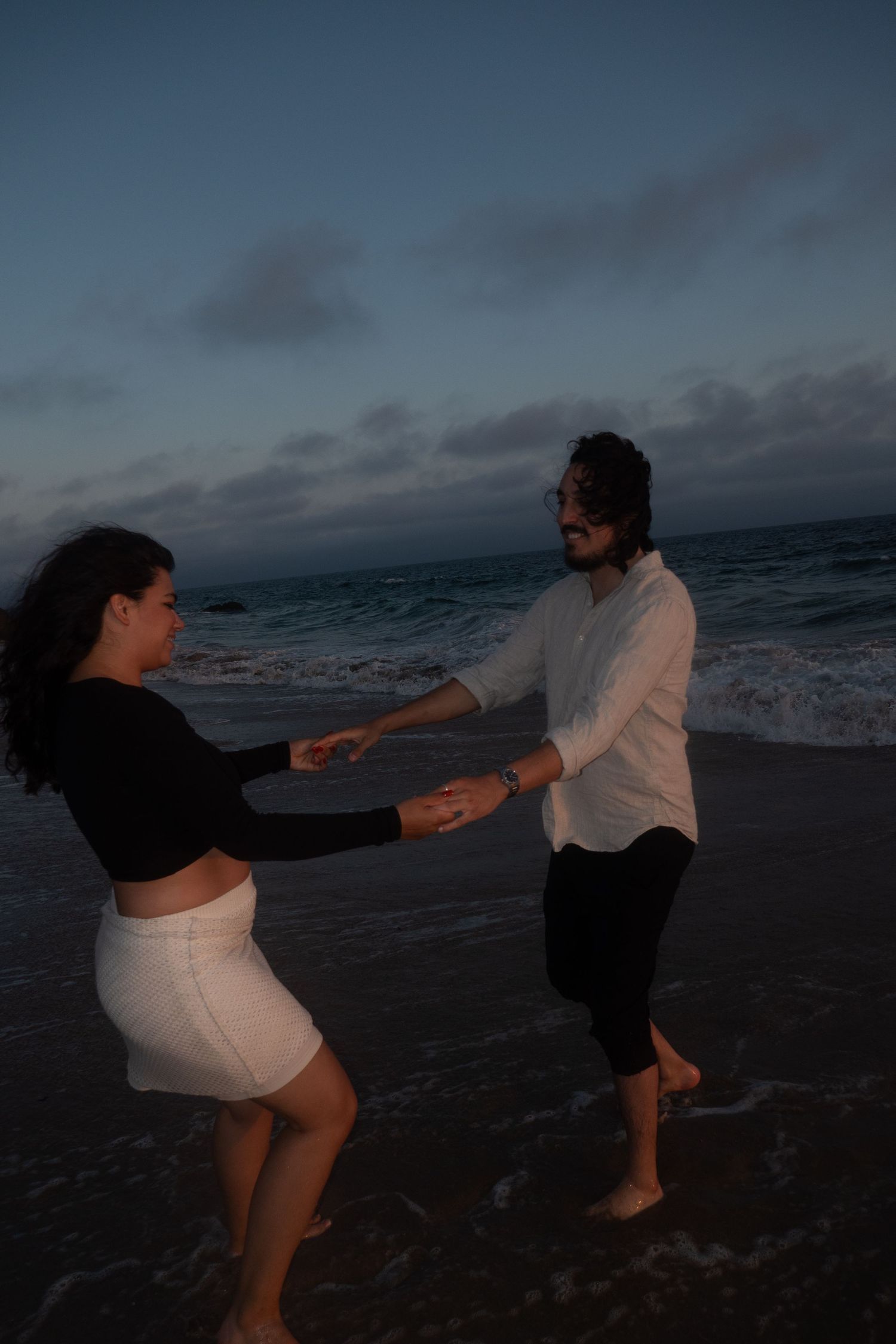 Silhouettes dance and play on a beach at dusk with waves crashing in the background.