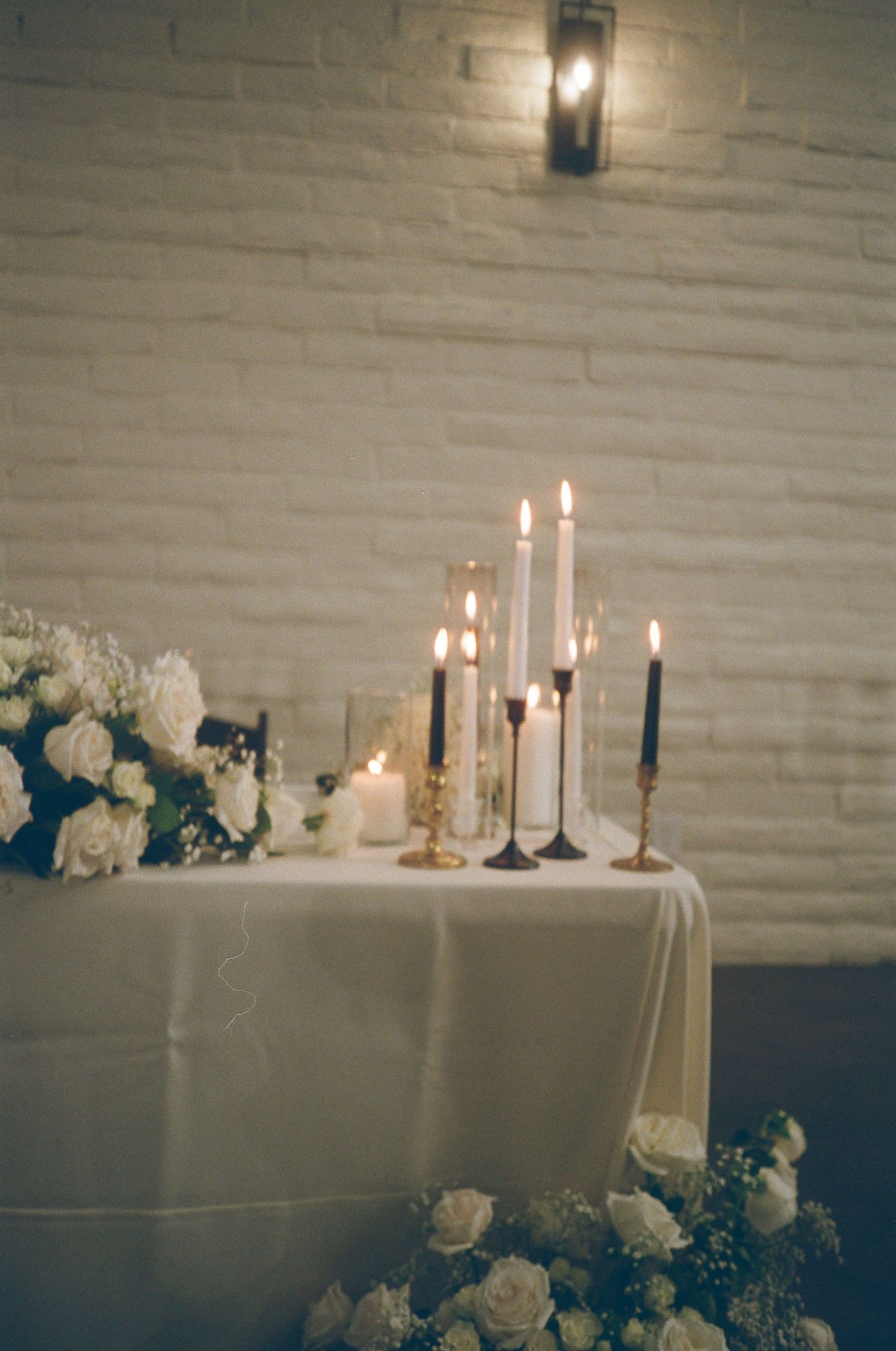 Romantic wedding sweetheart table with white roses and lit candles against white brick wall.