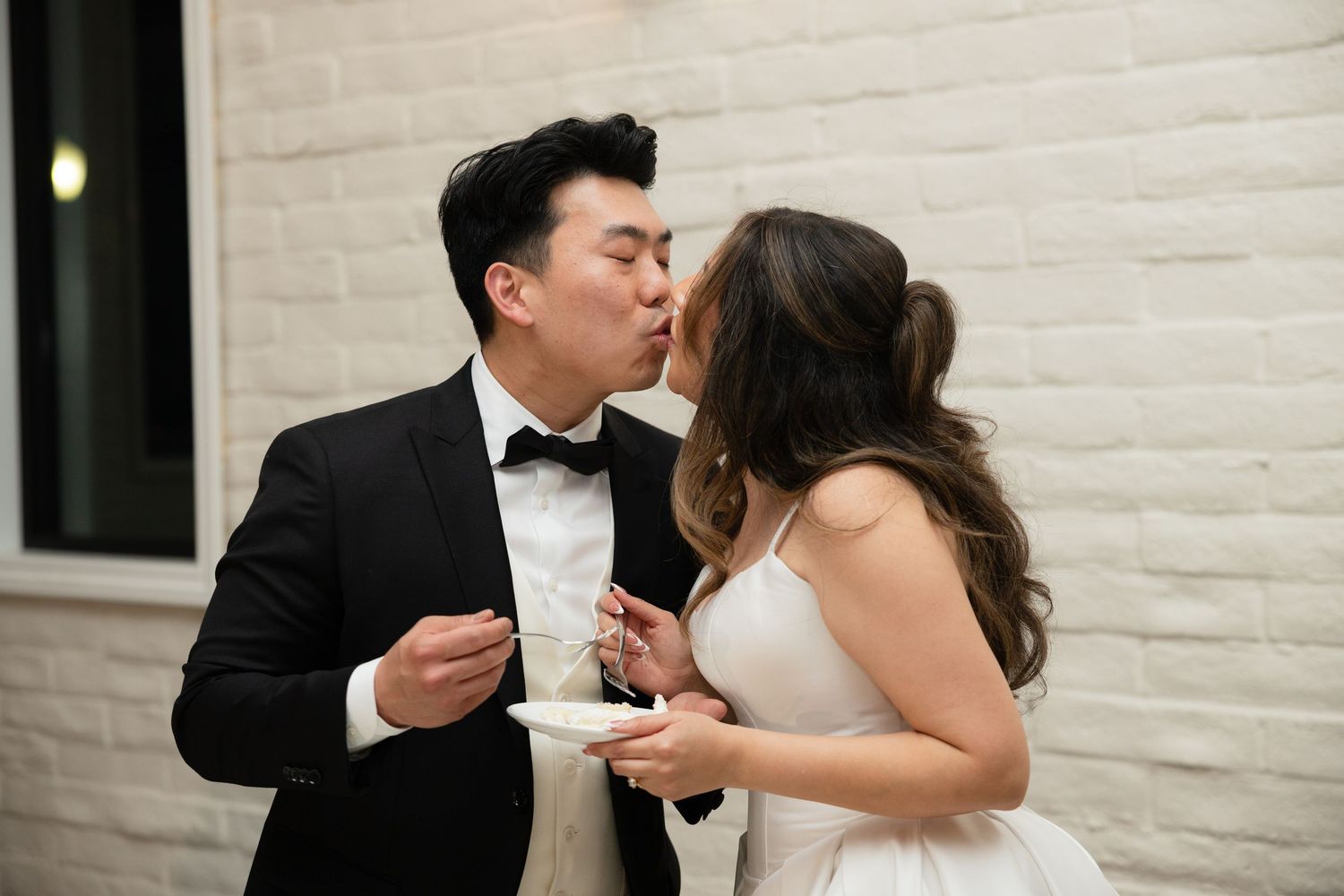 Bride and groom share a kiss while cutting wedding cake, bride in white dress groom in black tuxedo.