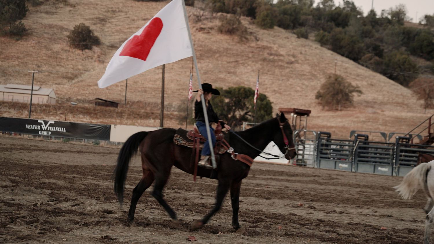 Rider on dark horse carrying white flag with red heart at outdoor rodeo arena.