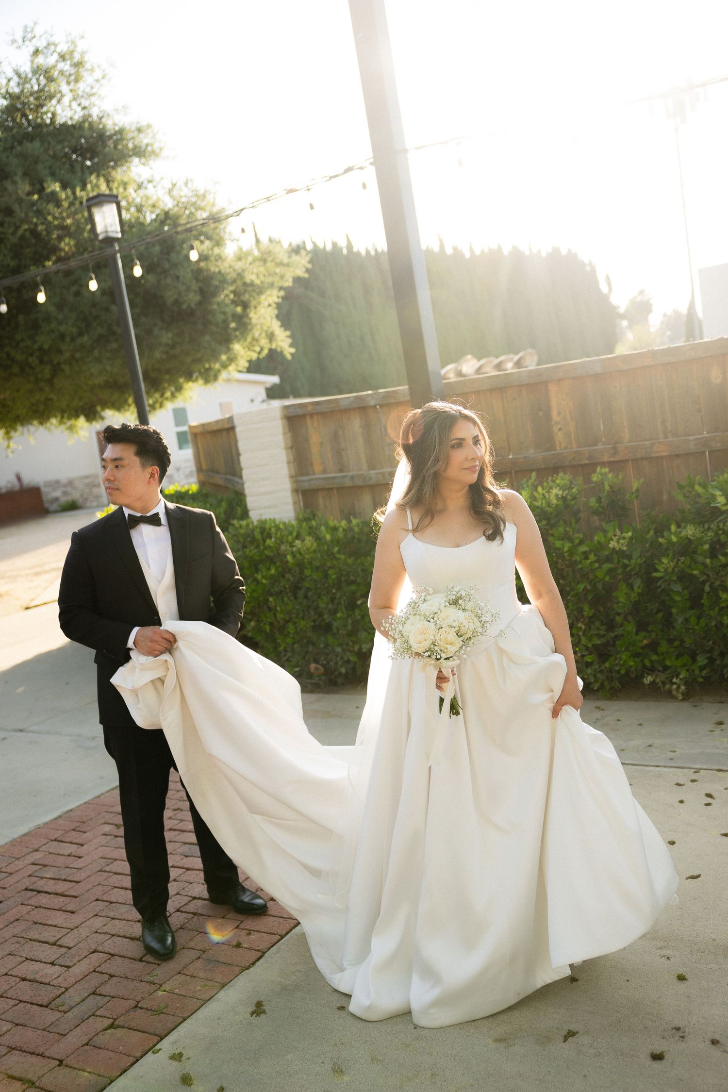 Bride in white ballgown holds bouquet as groom in tuxedo carries her train while walking outdoors in golden sunlight.