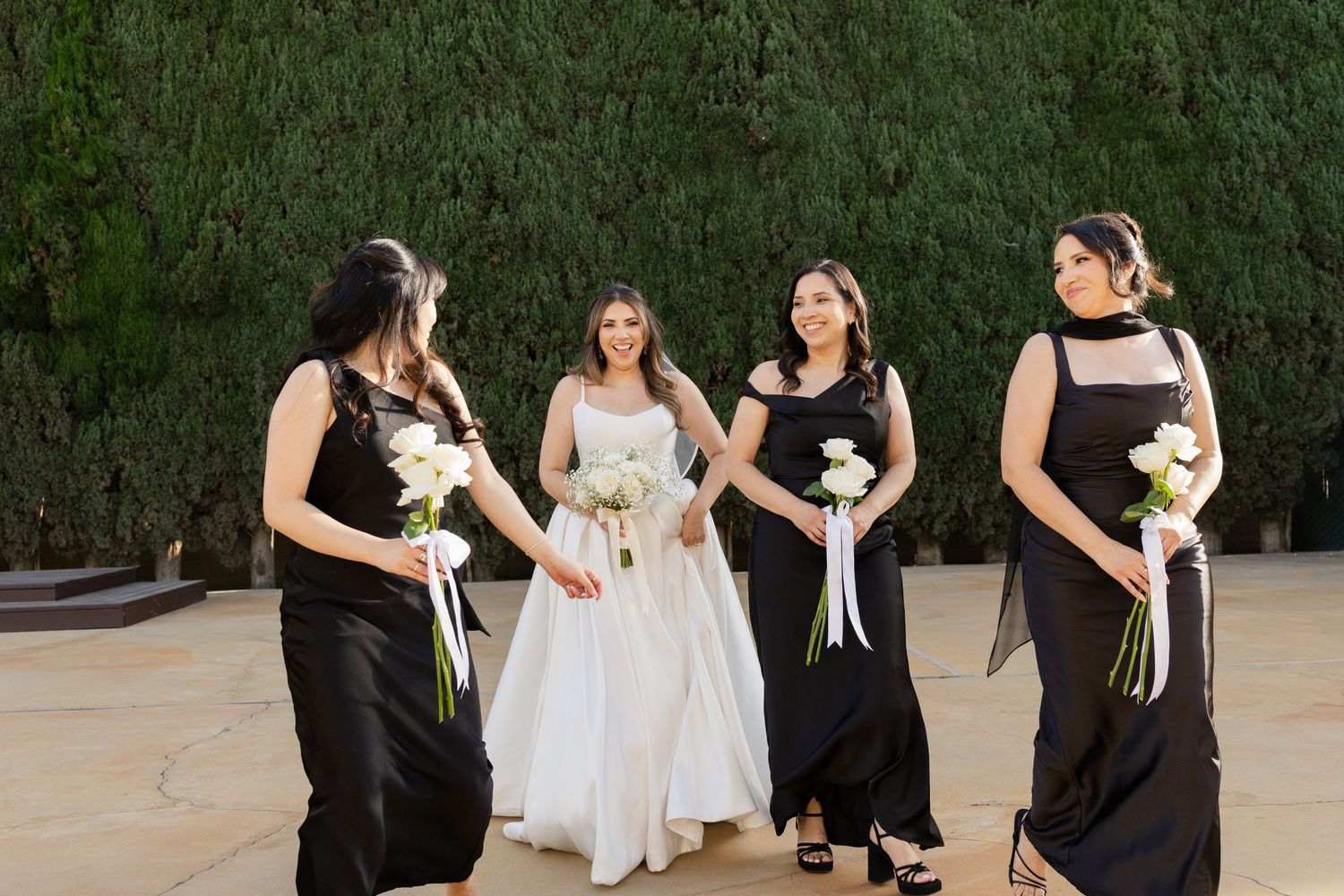 Bride in white gown with bridesmaids in black dresses holding white bouquets outdoors at wedding.