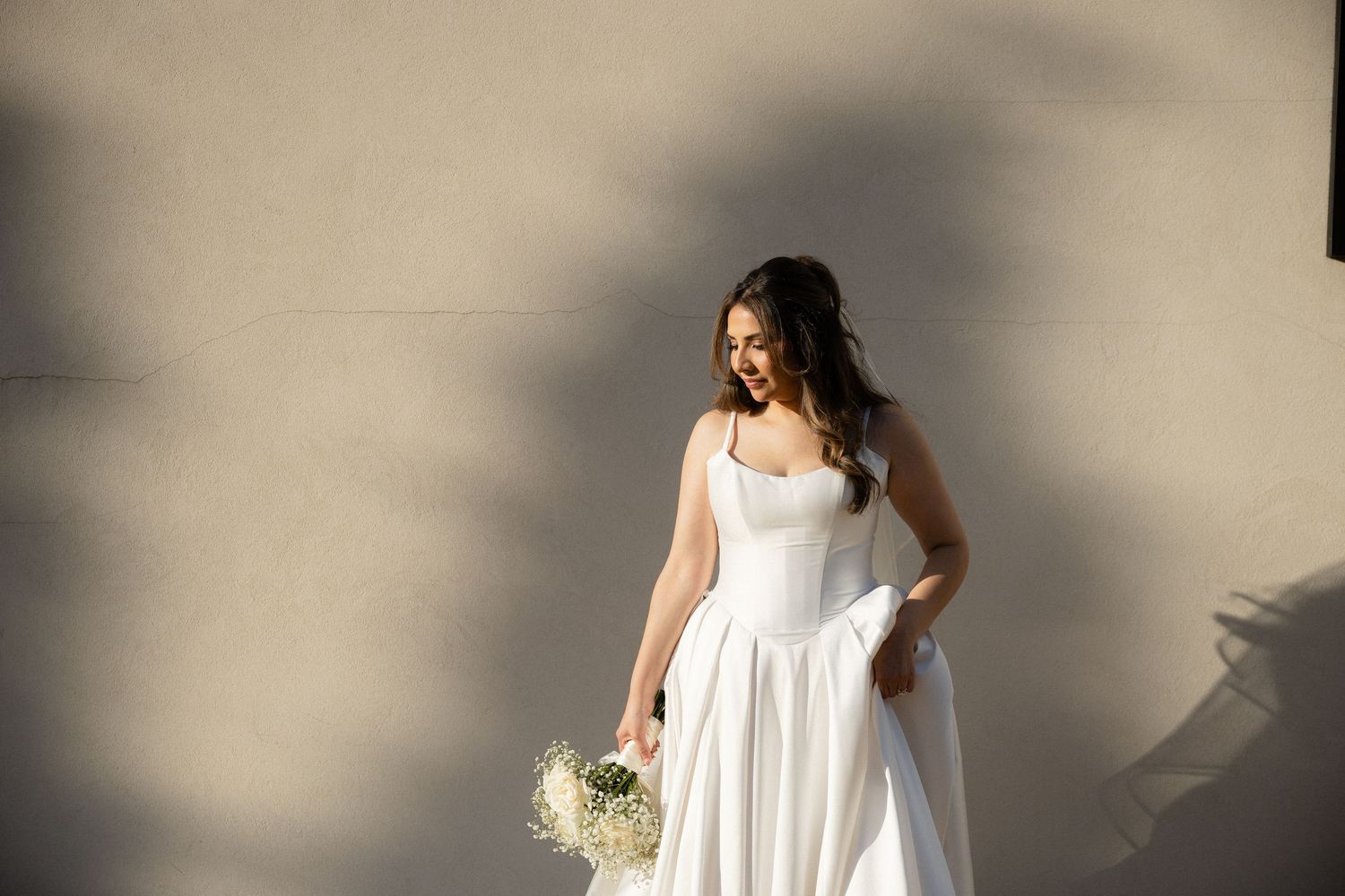 Bride in a white satin gown holding a bouquet, standing against a sunlit neutral wall.