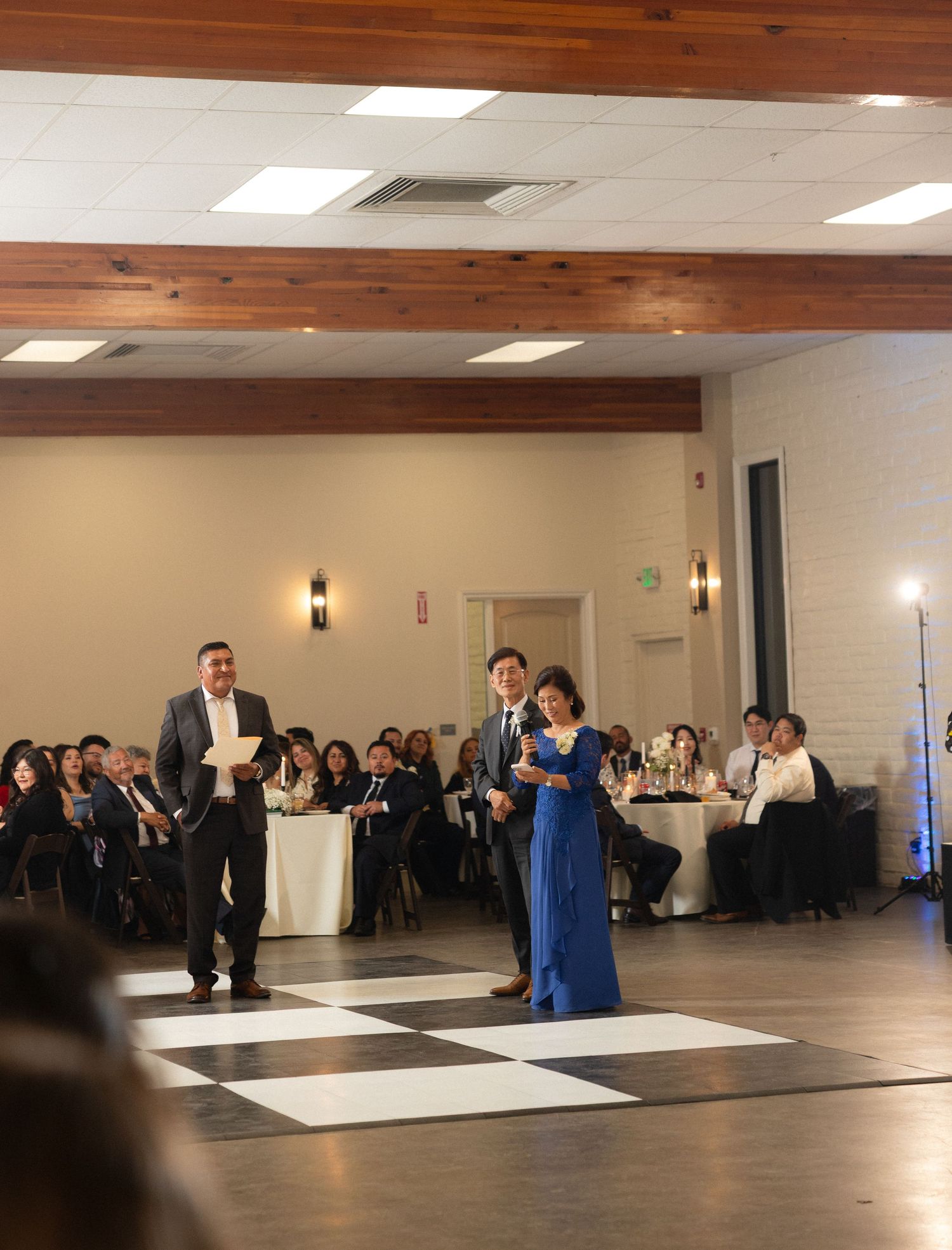 Man and woman in blue gown standing on checkered dance floor during wedding reception speech.