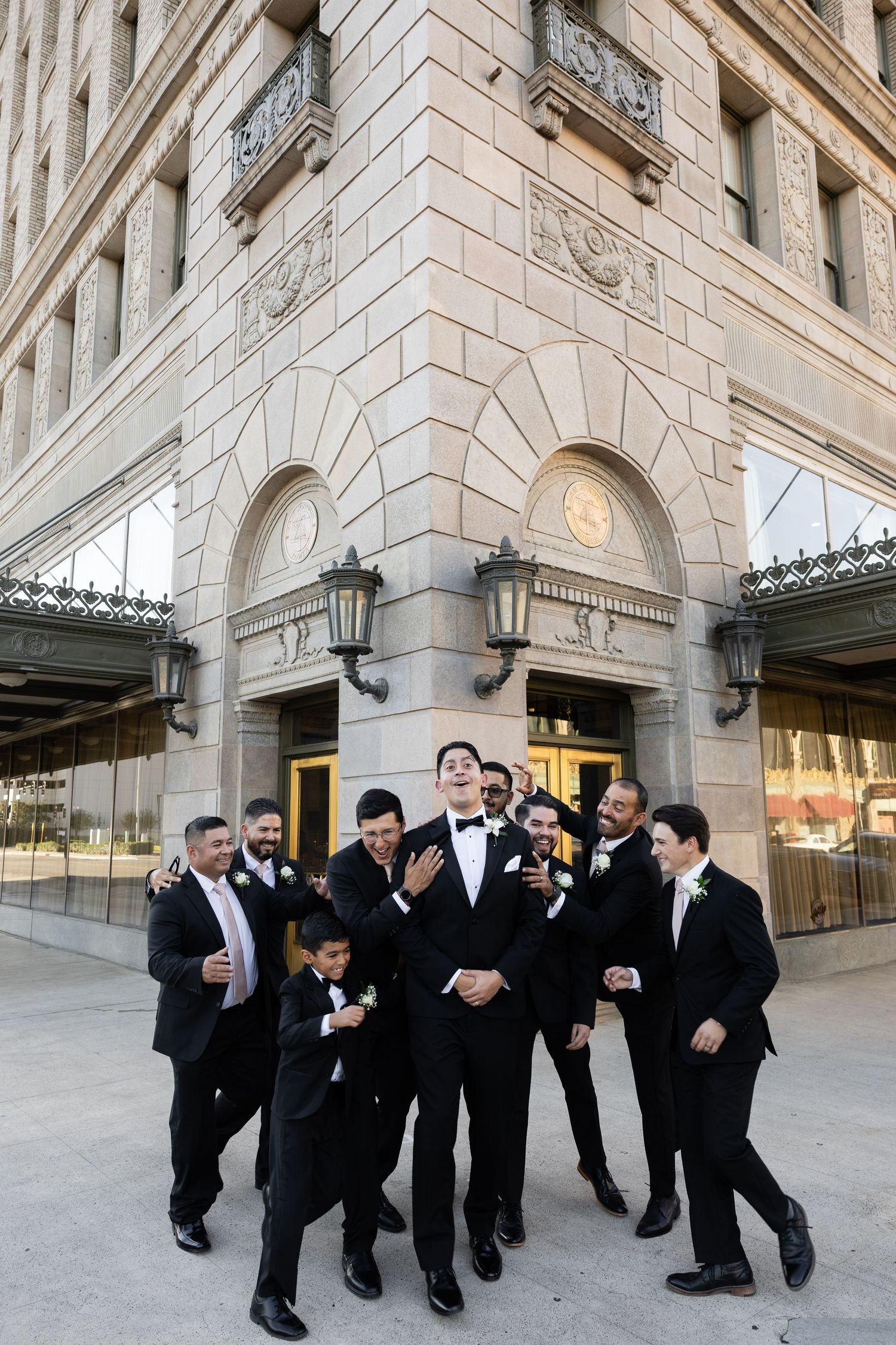 Groomsmen in black tuxedos having fun during photo session outside building.