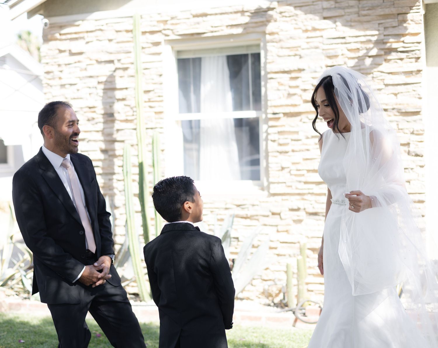 A young ring bearer in a black suit playfully tugs on a white wedding dress during a candid moment.