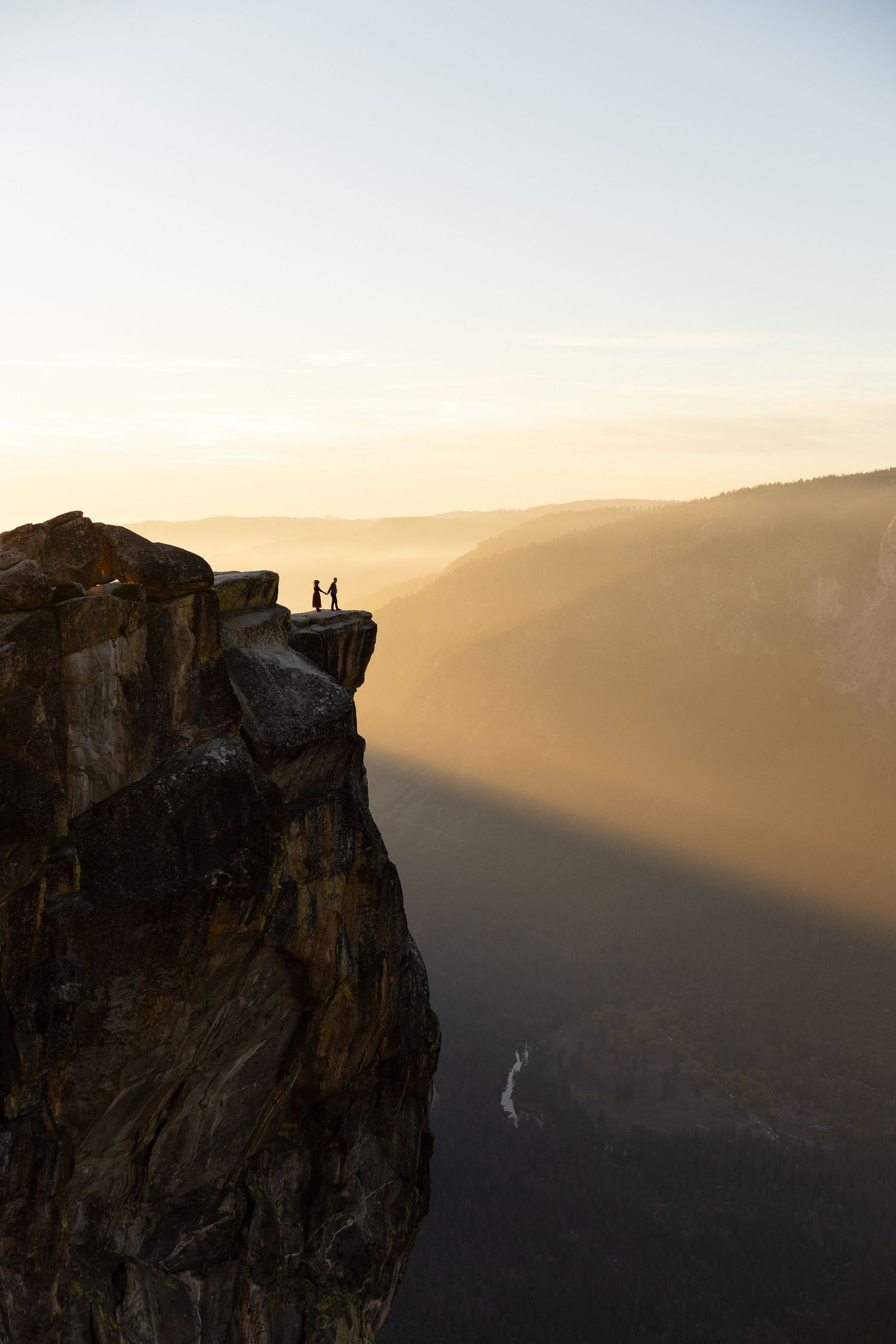 Two silhouettes stand on a dramatic cliff edge overlooking a misty valley at sunset.