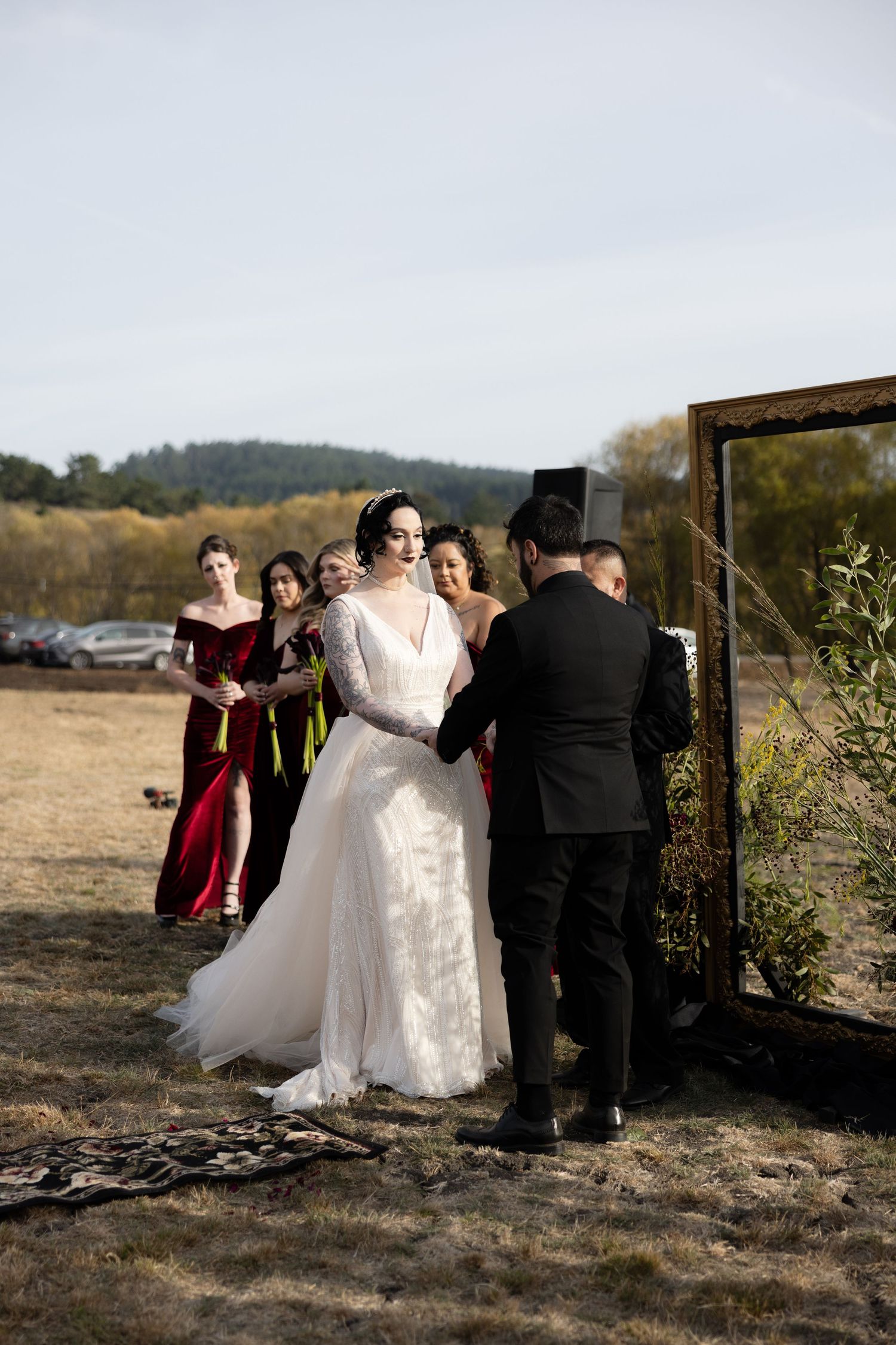 Wedding party gathers around a wooden arch frame during an outdoor ceremony in a field.