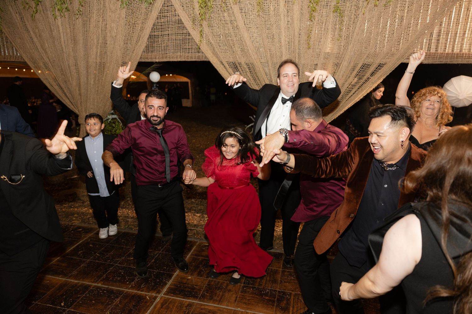 Lively group dance at wedding reception with guests in burgundy shirts jumping and celebrating.