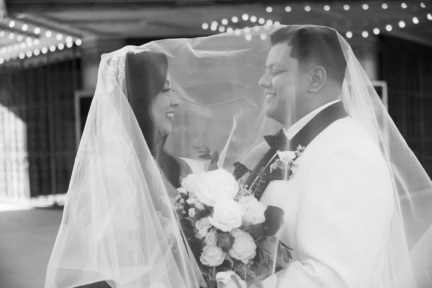 Black and white photo of bride and groom smiling under wedding veil, bride holding white bouquet.