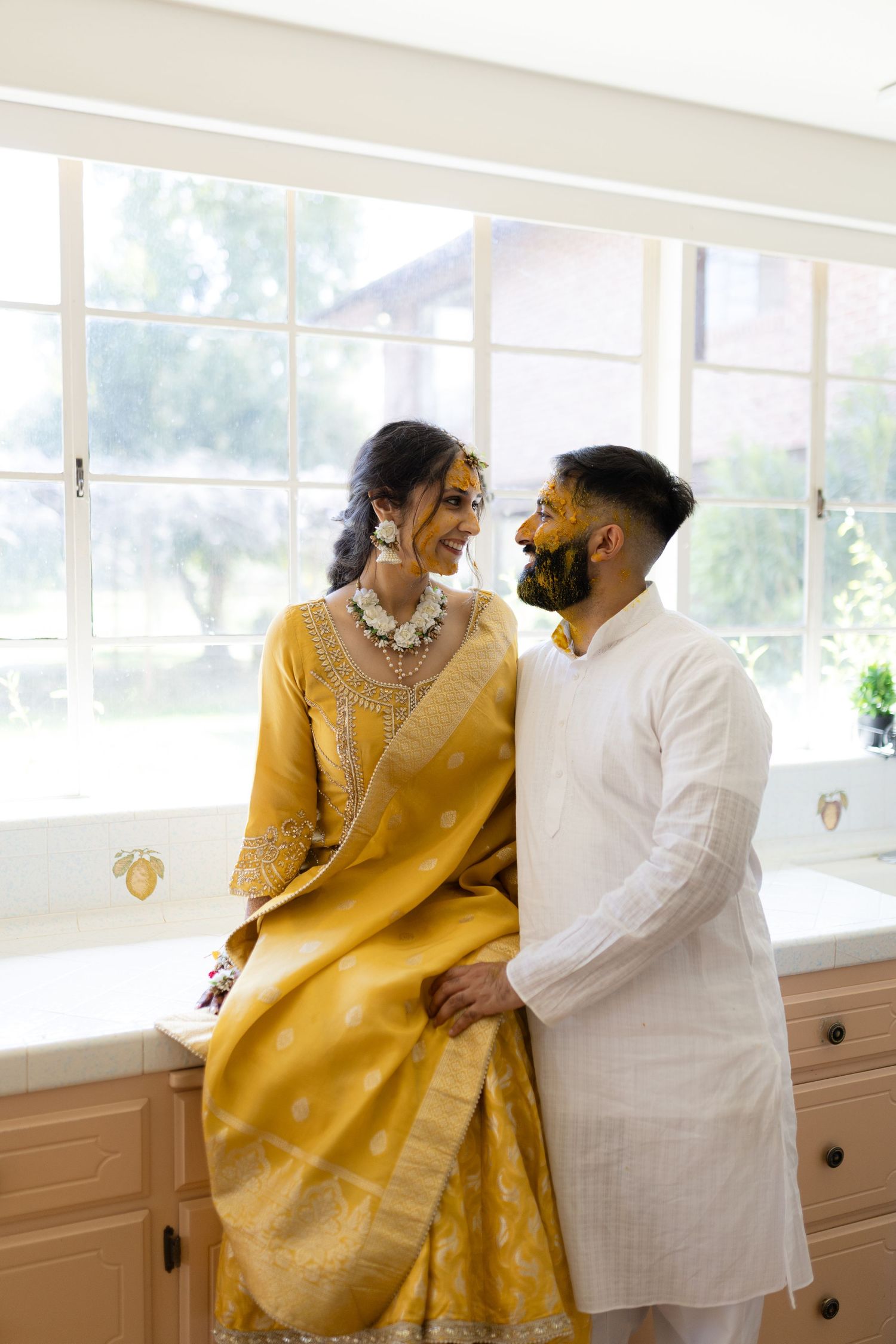 A couple in yellow and white formal attire sitting together by a bright window.