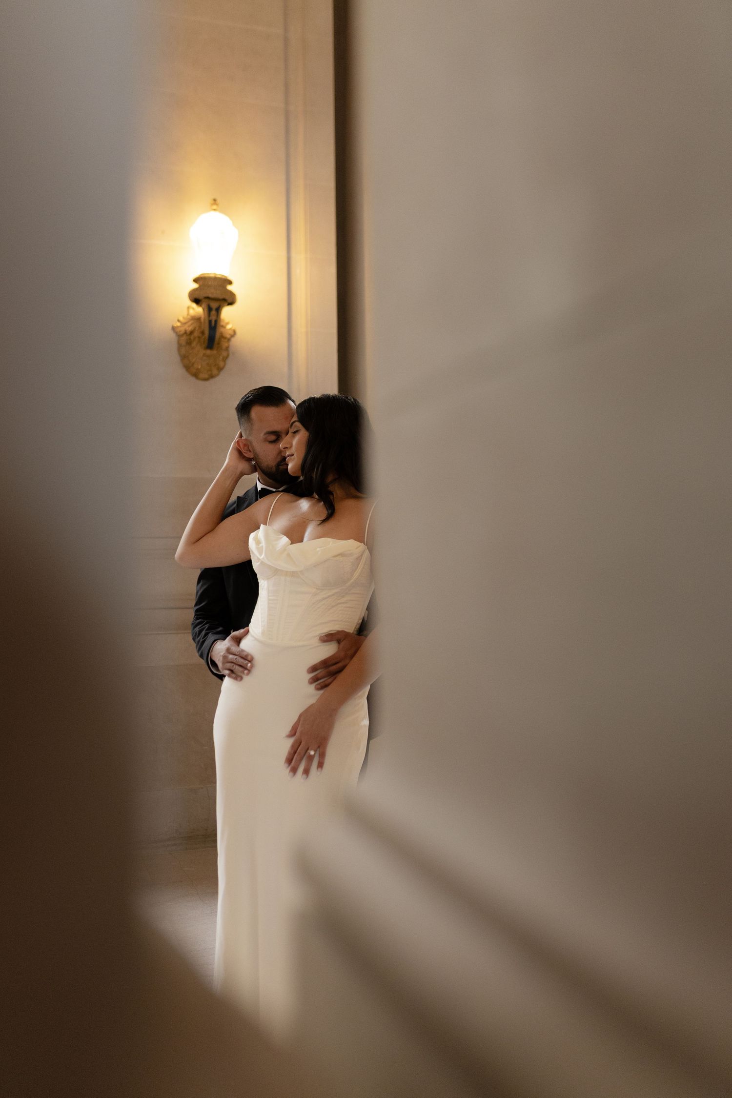 A romantic moment captured between a couple in formal attire sharing an intimate embrace near an illuminated wall sconce.