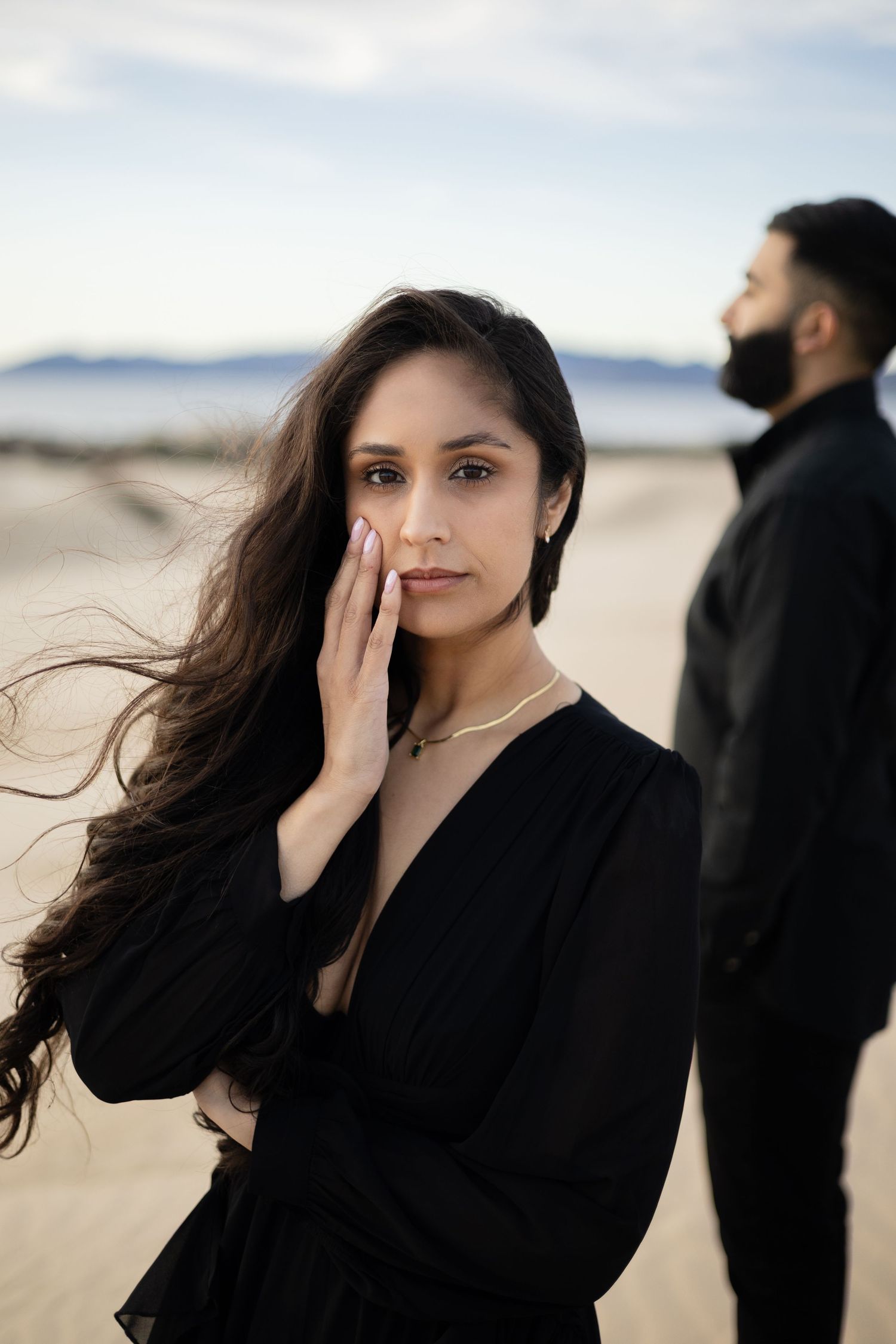 A woman in a black dress poses gracefully on a windy beach with mountains visible in the distance.