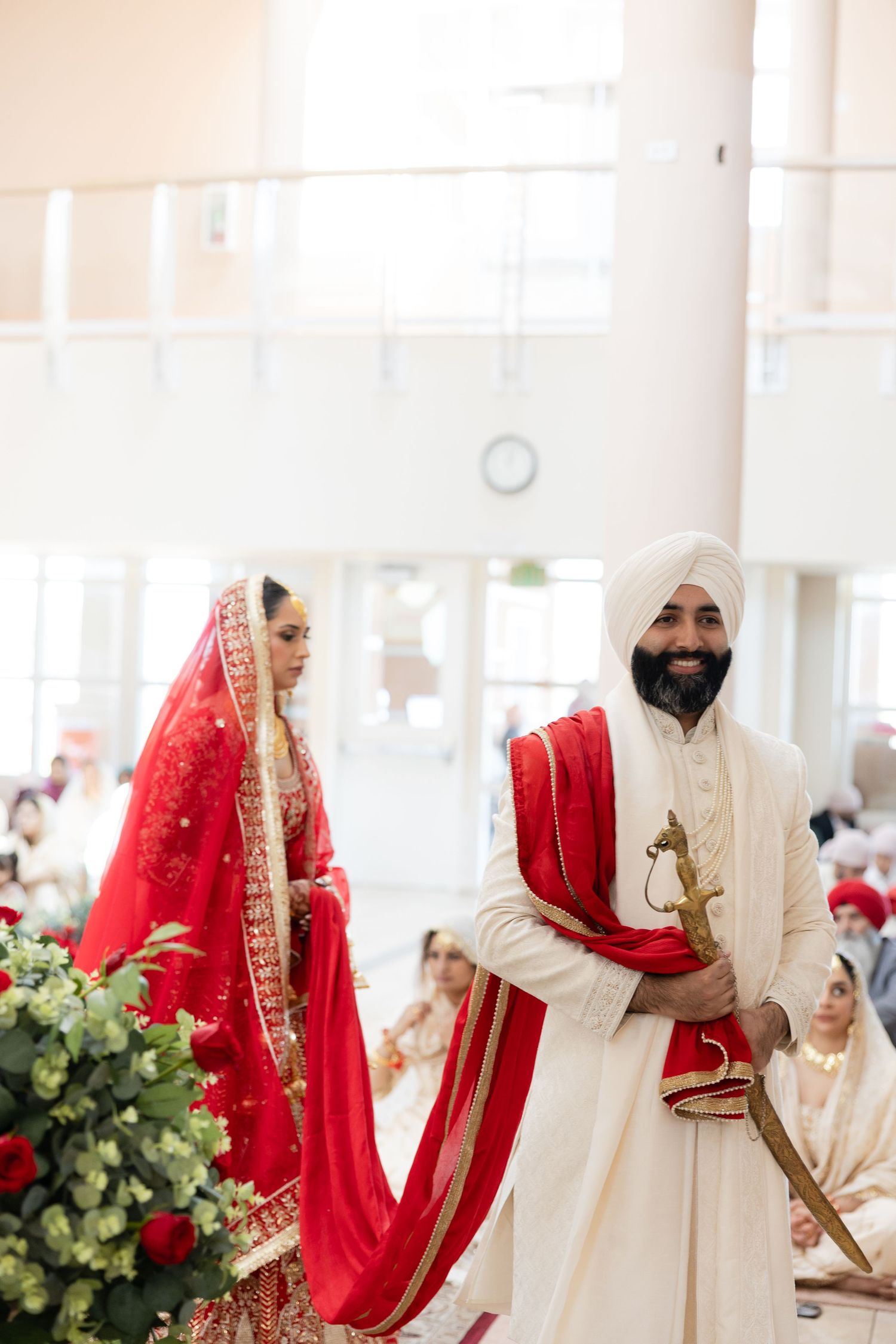 Couple in traditional Sikh wedding attire standing together with red and cream colored outfits.