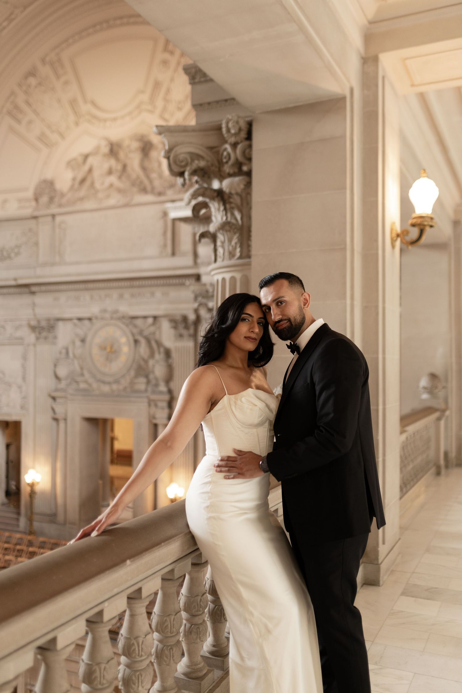 Elegant couple poses on ornate marble staircase in historic building with romantic lighting and architectural details.