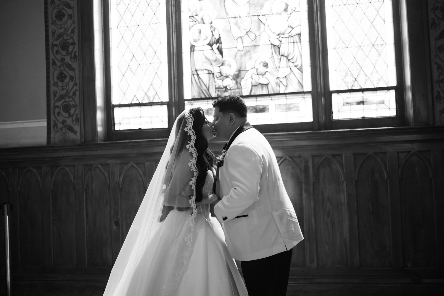 Black and white photo of a bride and groom sharing a kiss in a church with stained glass windows.