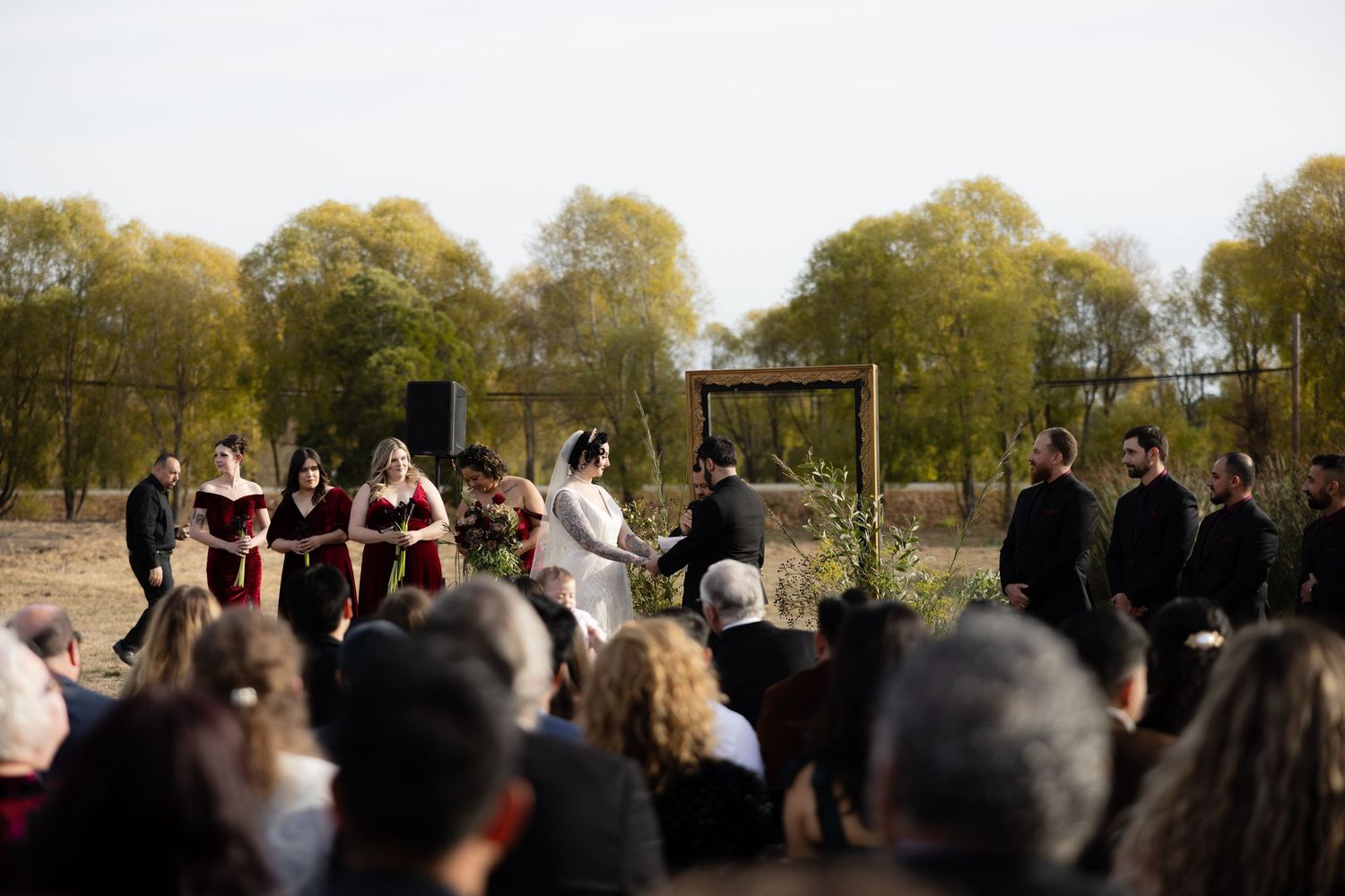 Guests watch outdoor wedding ceremony from rows of chairs in natural field setting.