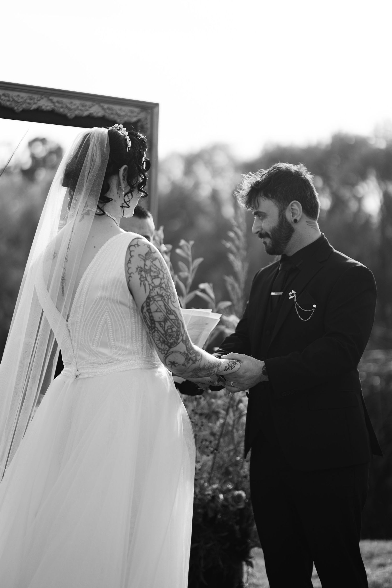 Black and white photo of a couple holding hands during their wedding ceremony under a wooden frame.