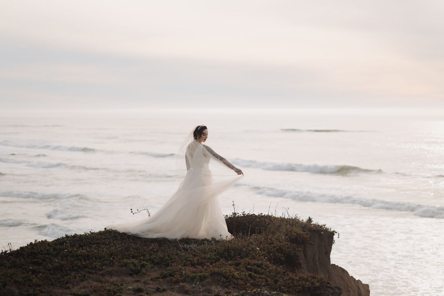 Bride in flowing gown dances on oceanside cliff during sunset.