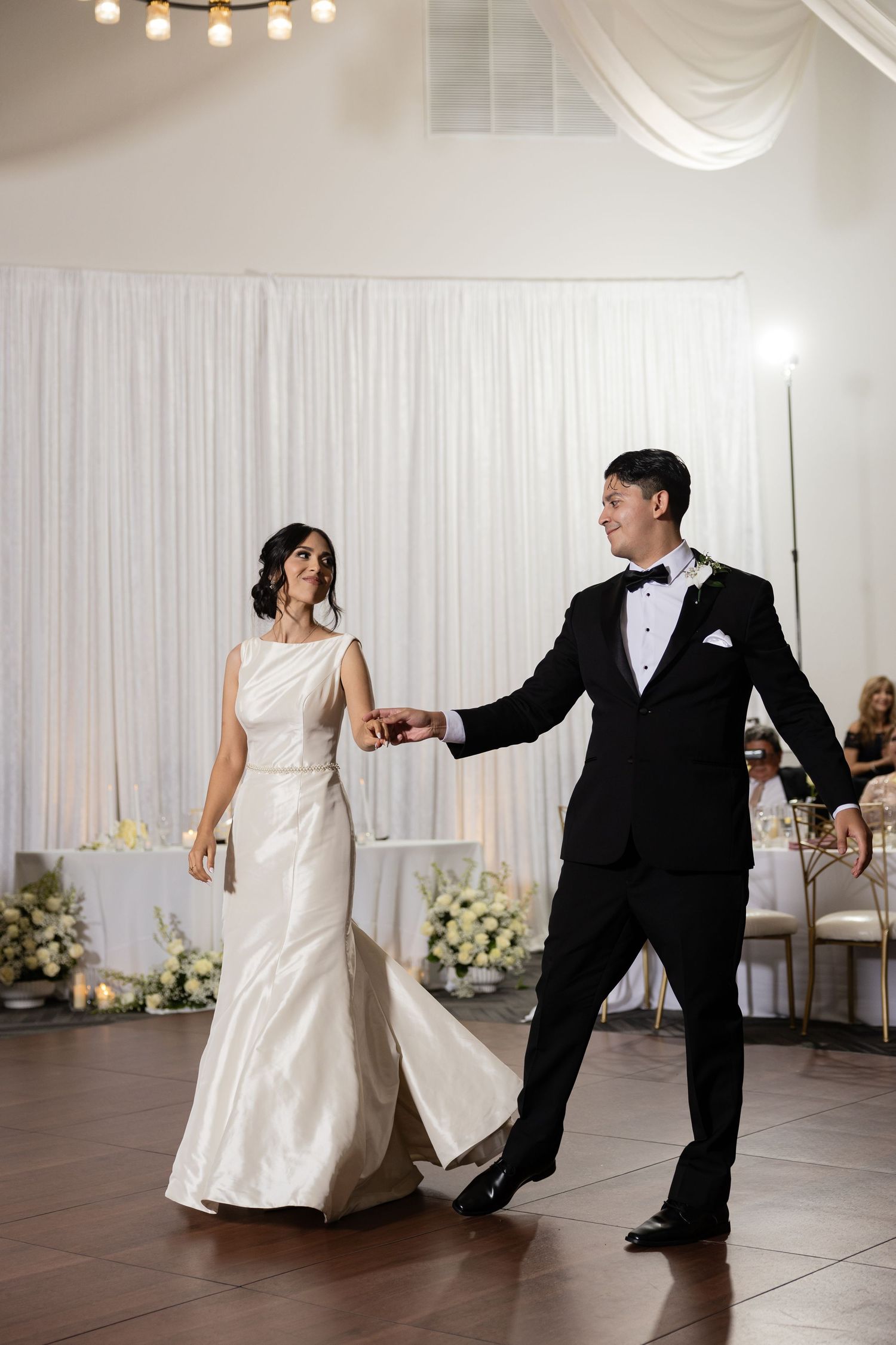 Wedding couple performs choreographed dance moves on wooden floor during reception celebration.