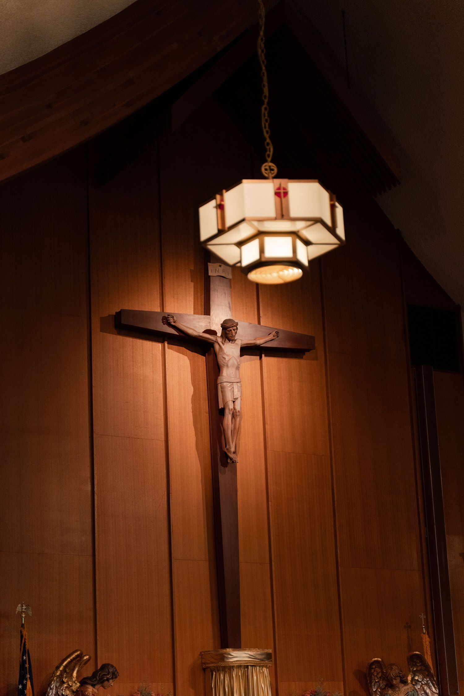 A wooden crucifix hangs beneath an art deco style pendant light in a church.