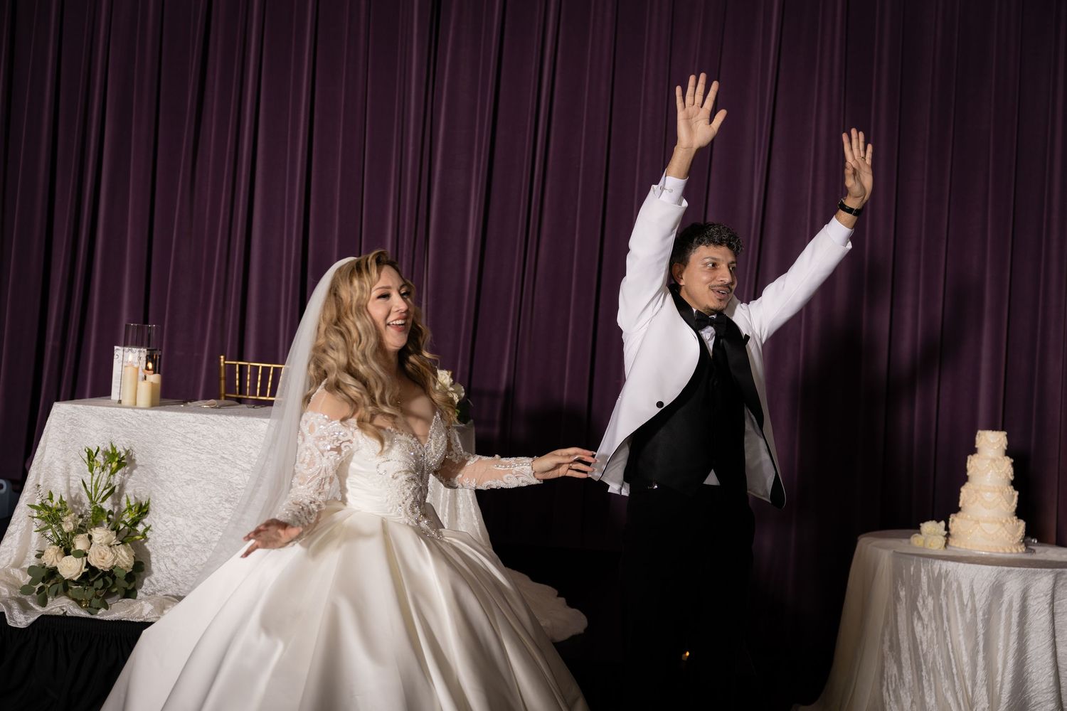 A couple celebrates energetically during their wedding reception with hands raised.