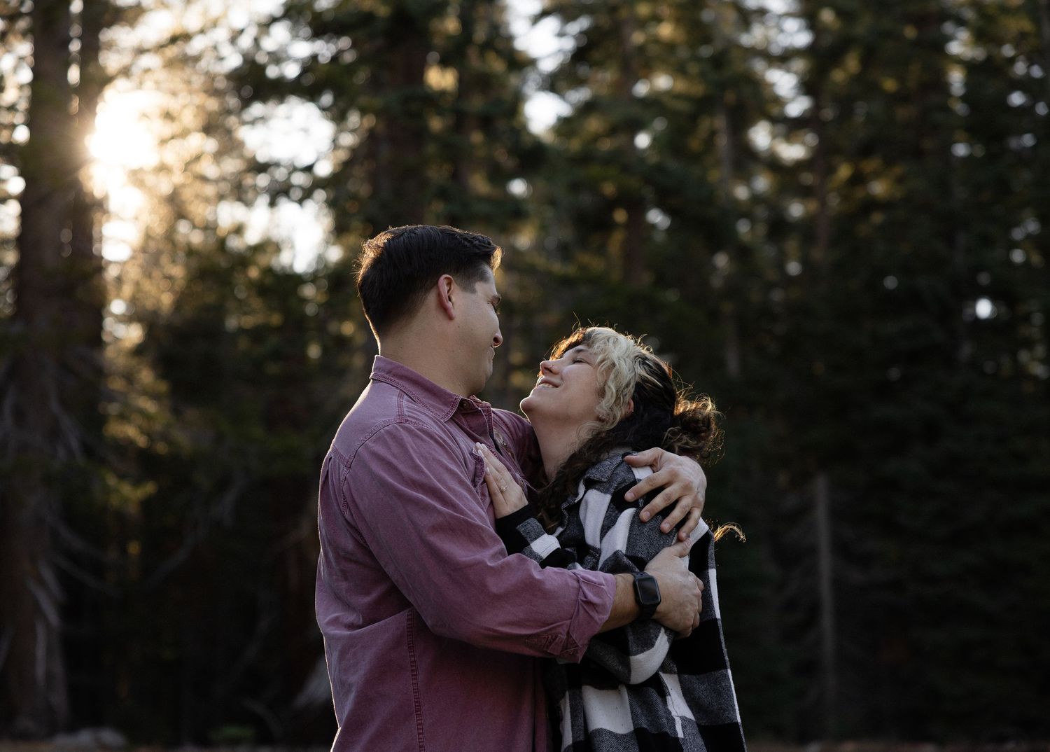 A tender embrace shared between two people against a backdrop of towering pine trees.