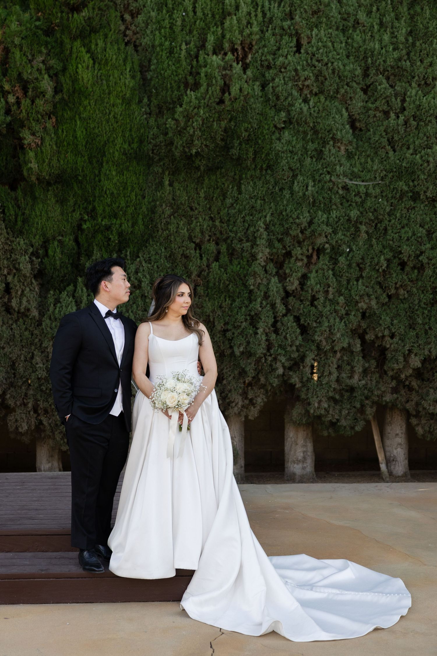 Bride and groom pose elegantly before lush green trees, she holds a white bouquet in a flowing satin gown.
