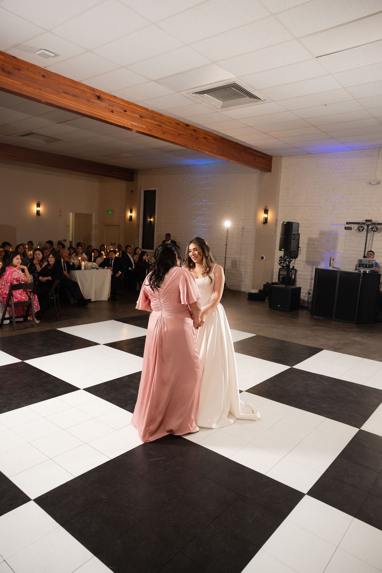 Bride and bridesmaid share an emotional dance on a black and white checkered floor at a wedding reception.
