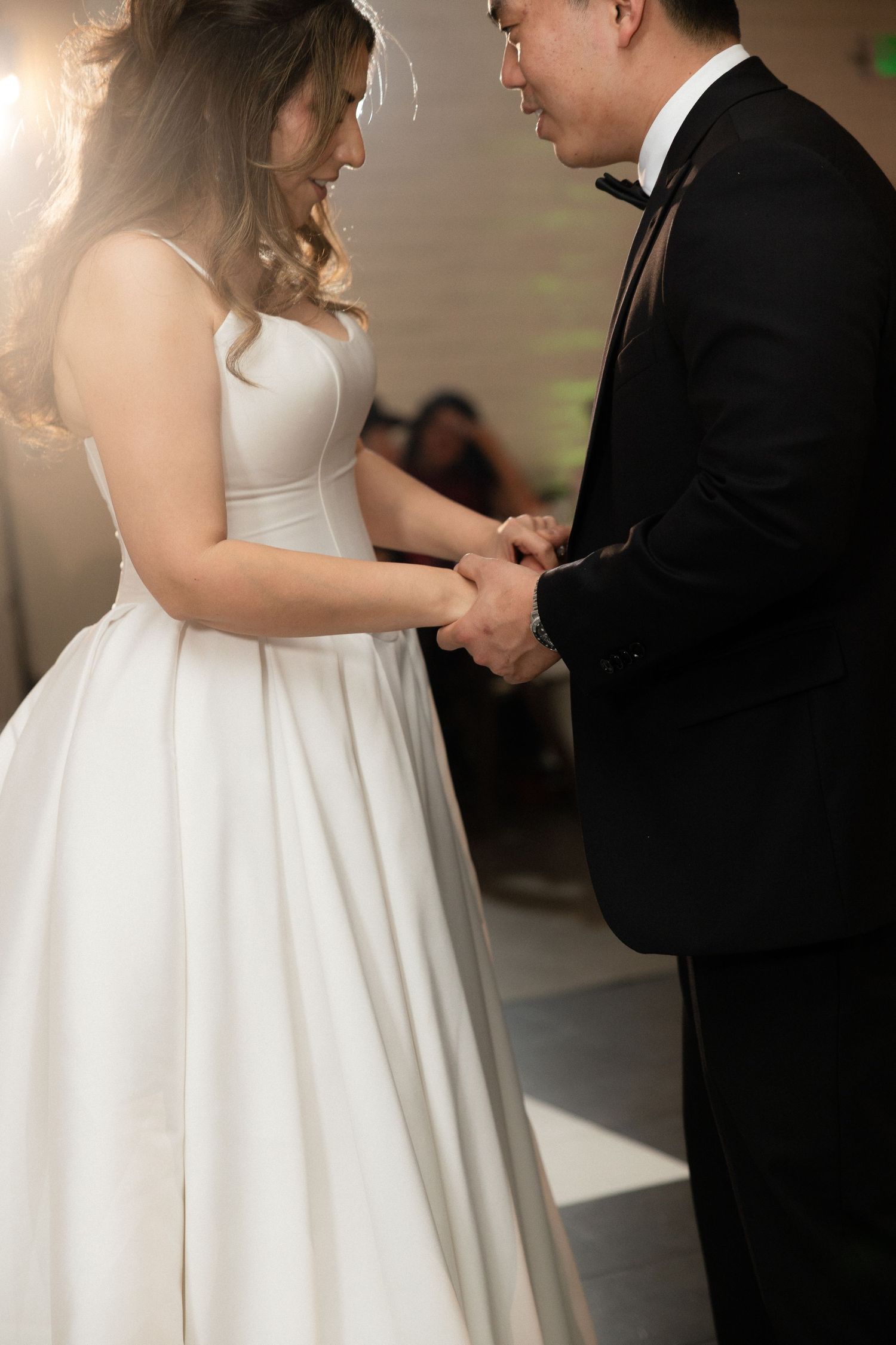 Bride in white ball gown holds hands with groom in black tuxedo during first dance ceremony.