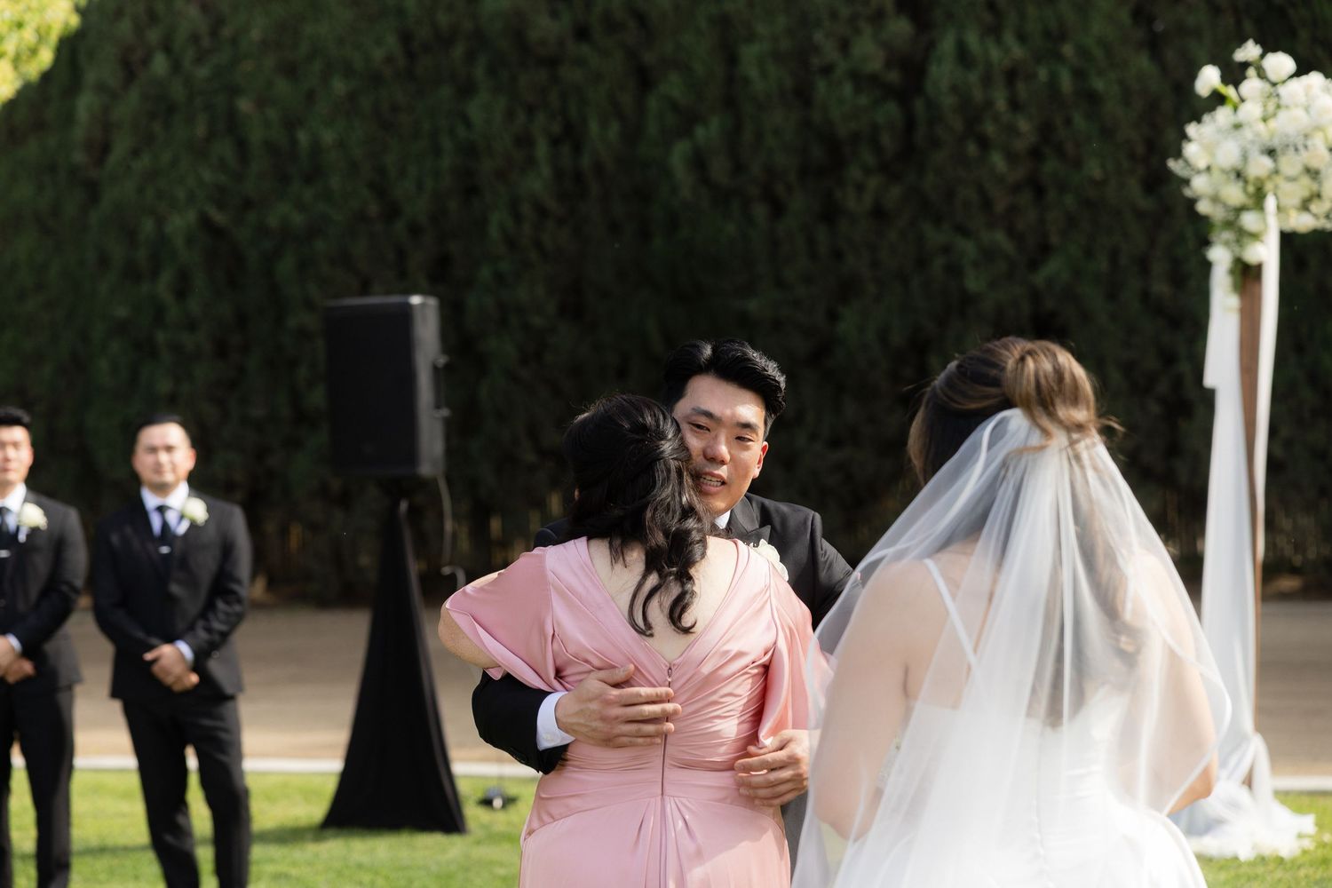 Groom embraces family member in pink dress as bride in white veil stands nearby outdoors.