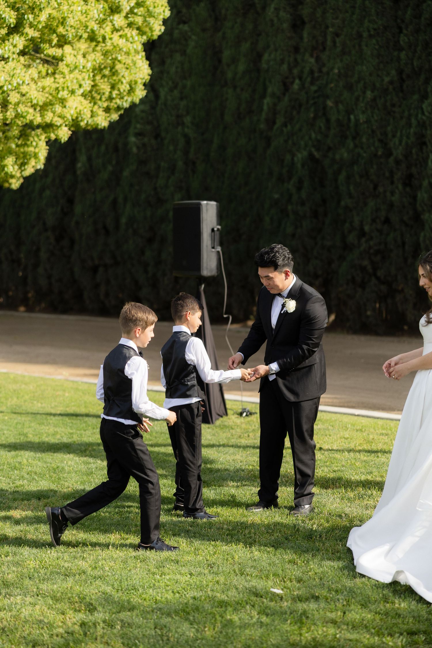 Groom in black tuxedo receiving wedding rings from young ring bearers during outdoor garden ceremony.