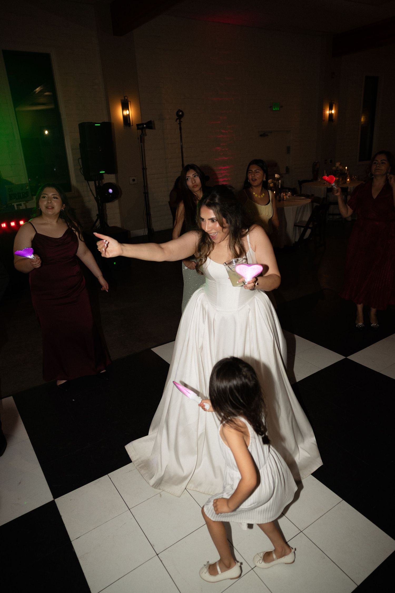 Bride dances with young flower girl on checkered dance floor surrounded by bridesmaids at wedding reception.