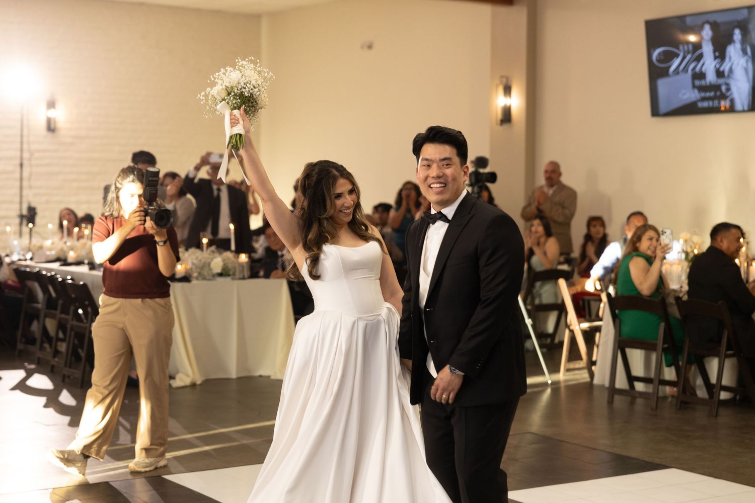 Joyful newlyweds make grand entrance on black and white checkered dance floor as bride raises bouquet to cheering wedding guests.